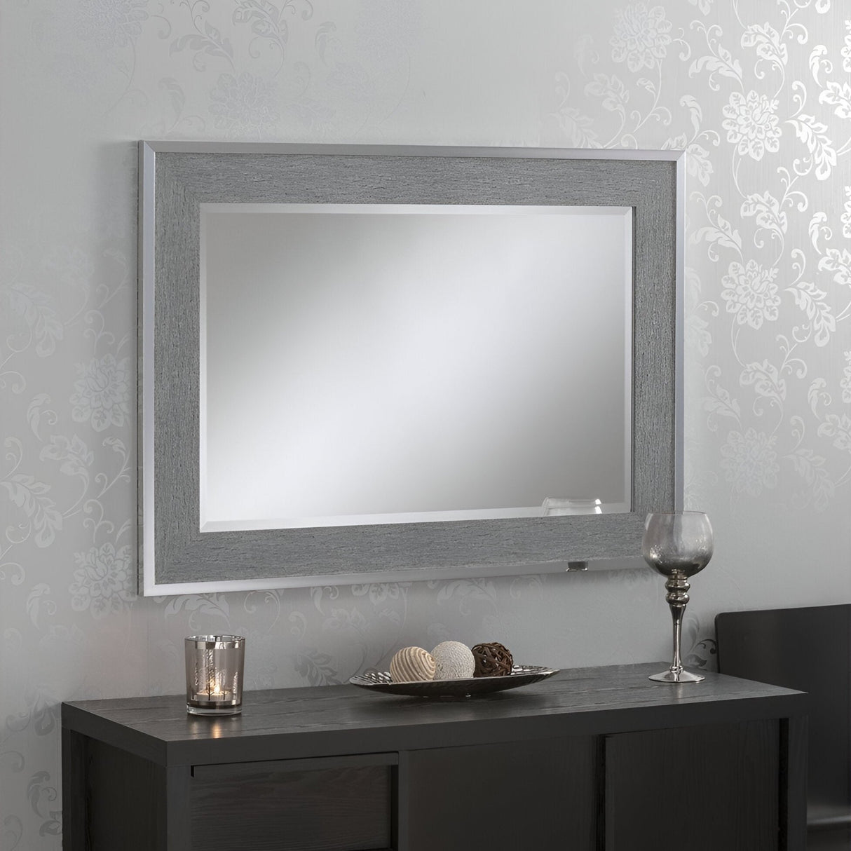 The Zennia Rectangle Wall Mirror - Silver is elegantly displayed on a patterned light-gray wall. Below, a dark console table enhances the contemporary decor, adorned with a glass candle holder, a decorative bowl filled with spheres, and an empty wine glass.