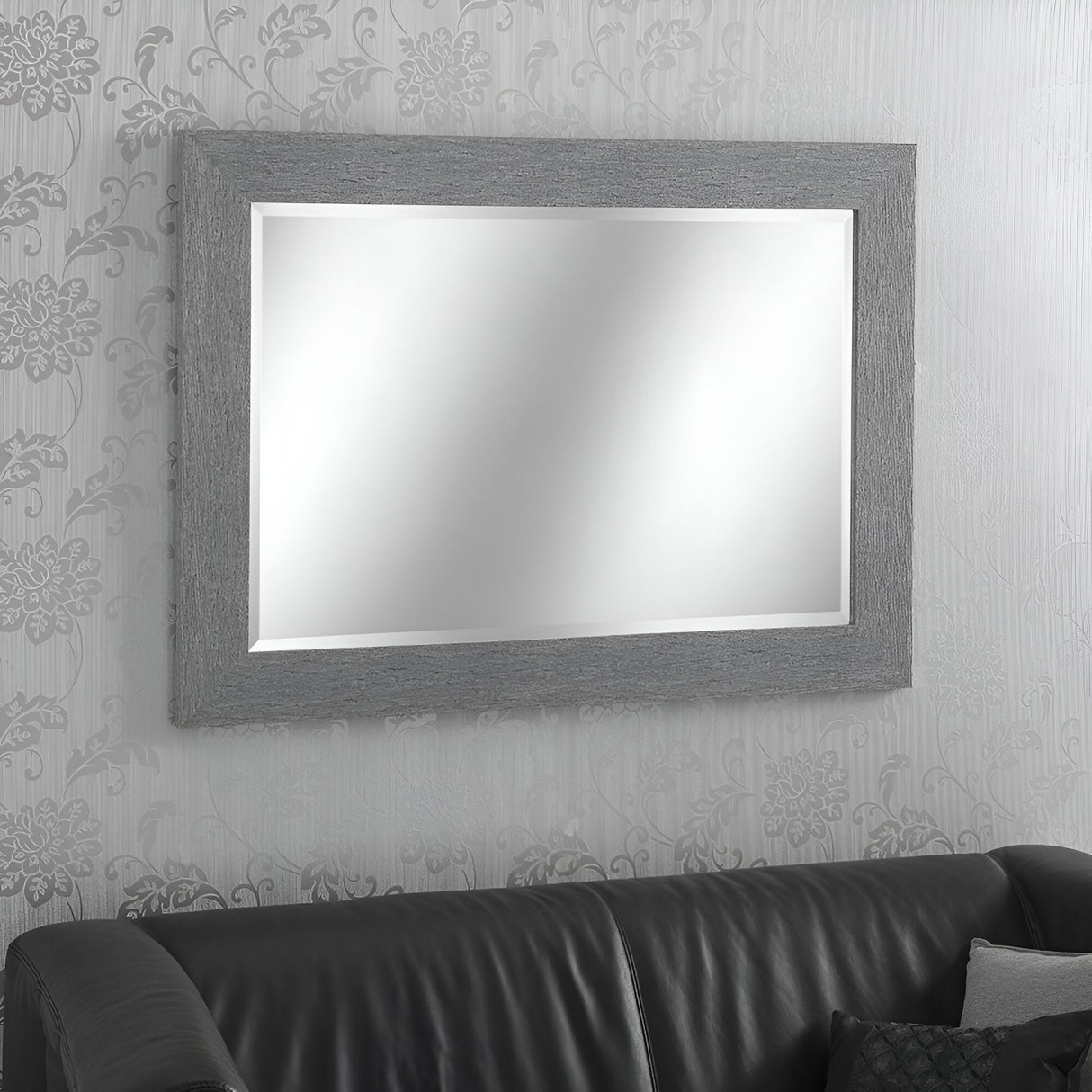 A Zennia Rectangle Wall Mirror - Grey is elegantly displayed on a wall decorated with floral-patterned wallpaper. Below this modern home accessory is a black leather sofa, accentuated by a dark cushion placed in the corner.
