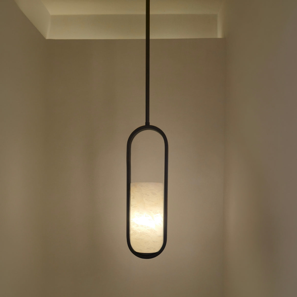 Eliona Capsule Alabaster Pendant Light in matt black, a single pendant with cylindrical alabaster shade held within an oval black frame, shown illuminated in a hallway setting.