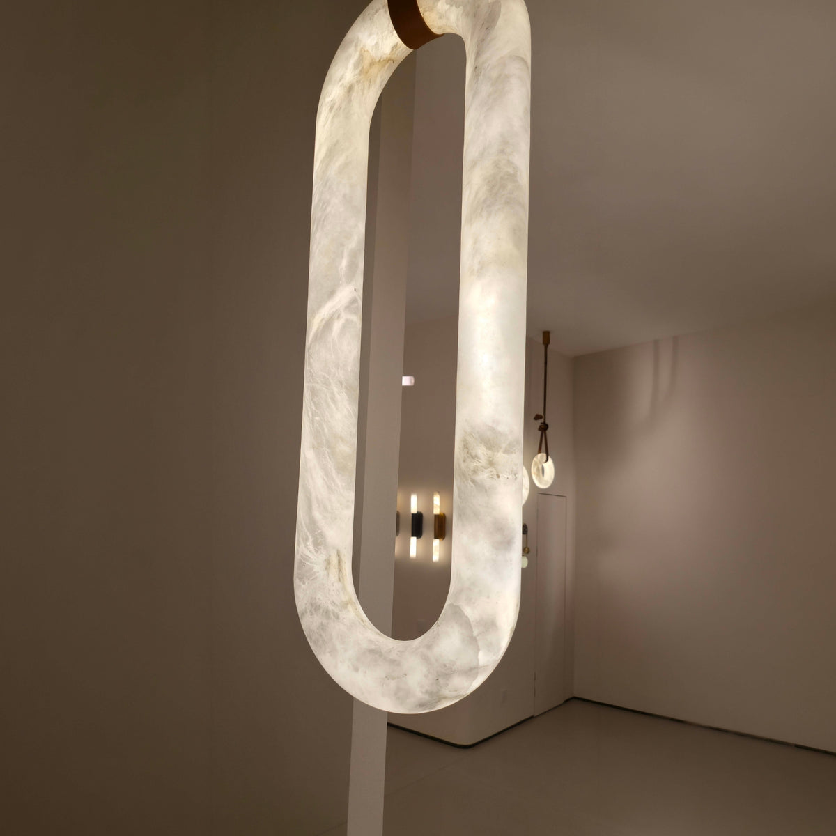 Serava Large LED Alabaster Pendant Light in Soft White, elongated capsule form with copper leather strap detail, glowing warmly in a modern living room setting.