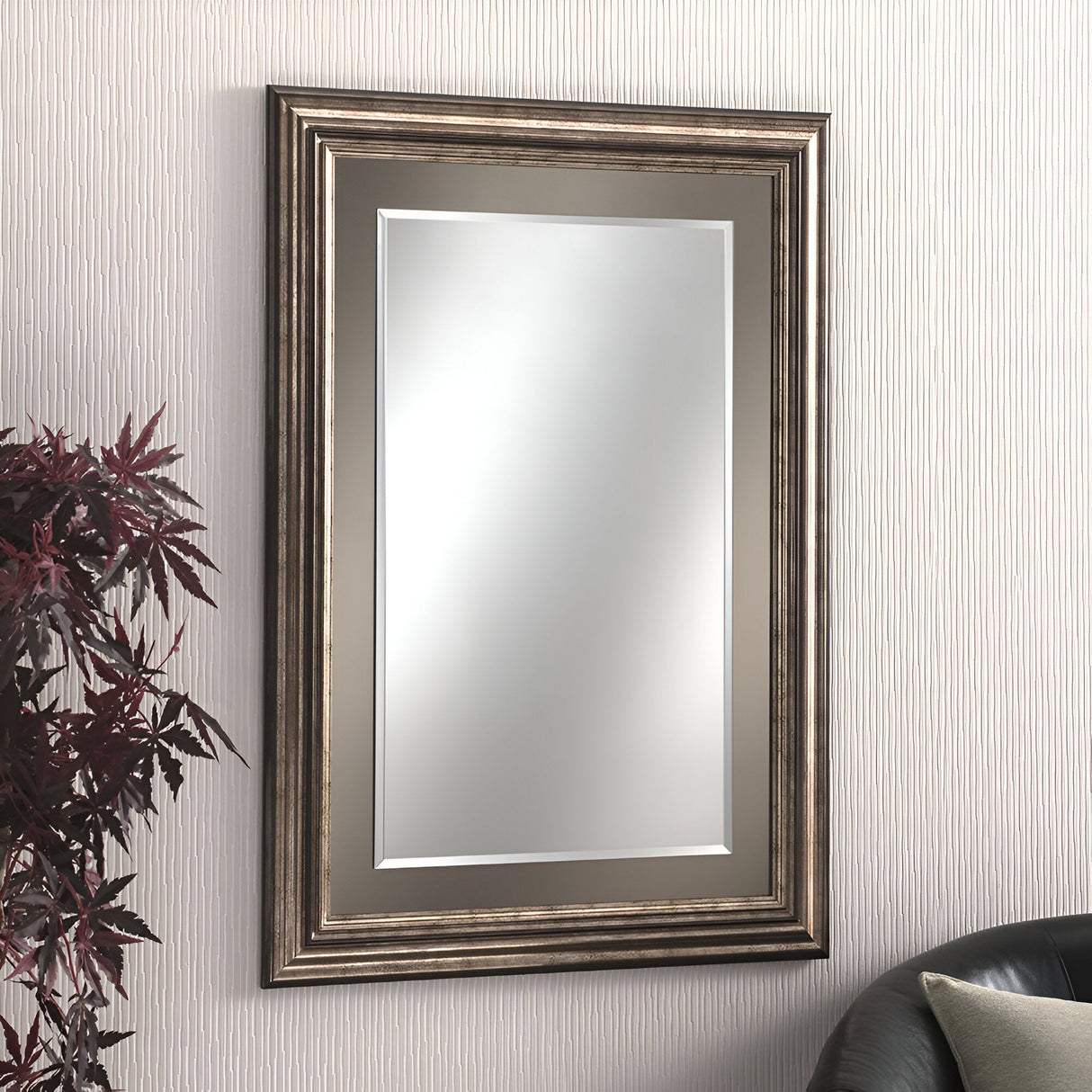 The Serenade Rectangle Wall Mirror - Bronze Lacquered, featuring a decorative wooden frame, elegantly adorns a textured white wall. A plant with dark leaves peeks in on the left, while a black couch with a green cushion completes this harmonious home decor scene.