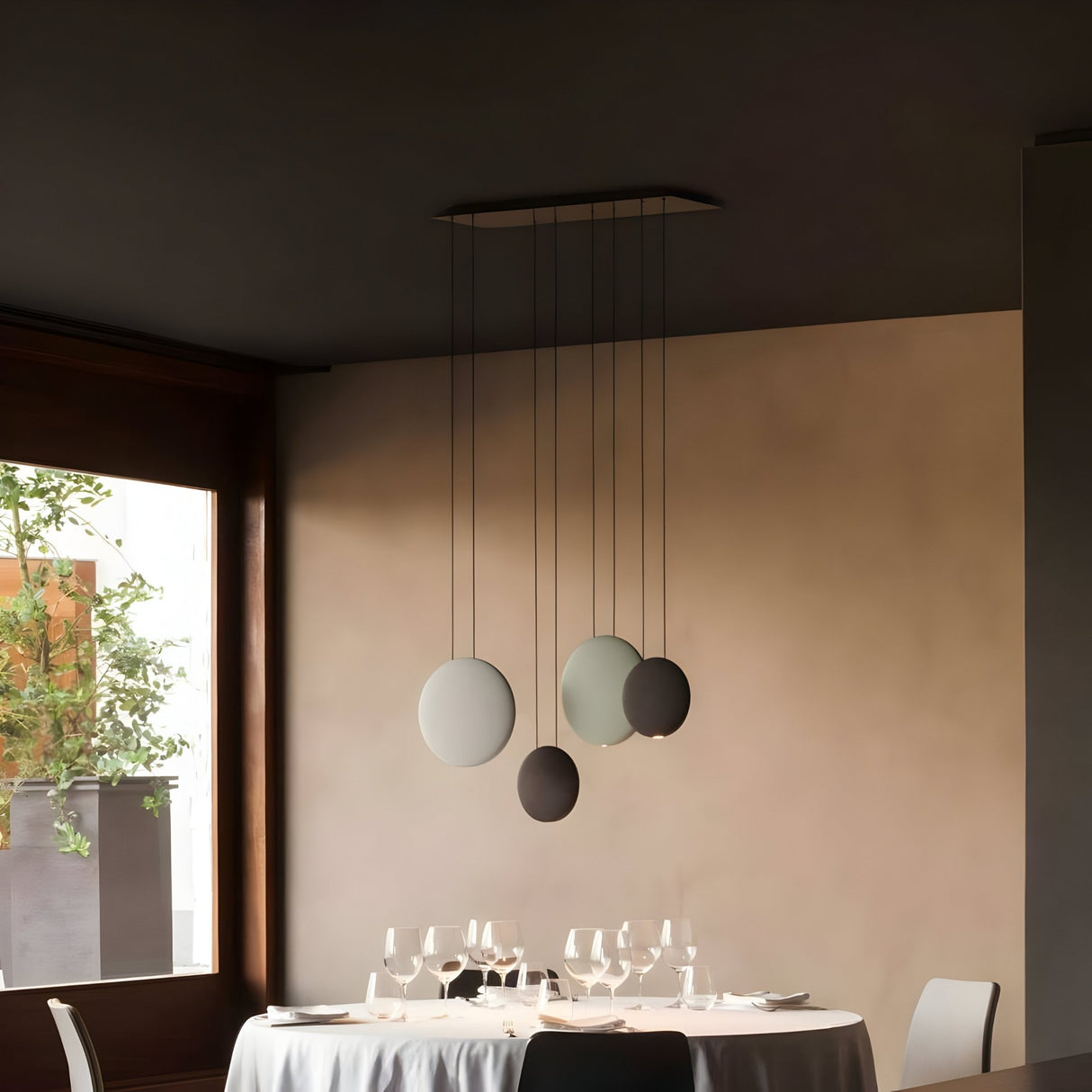 A modern dining area with a round table set for four, decorated with a white tablecloth, glasses, and plates. Four Cosmos LED Large Pendant Lights offer ambient lighting as sunlight streams through the window with lush greenery outside.