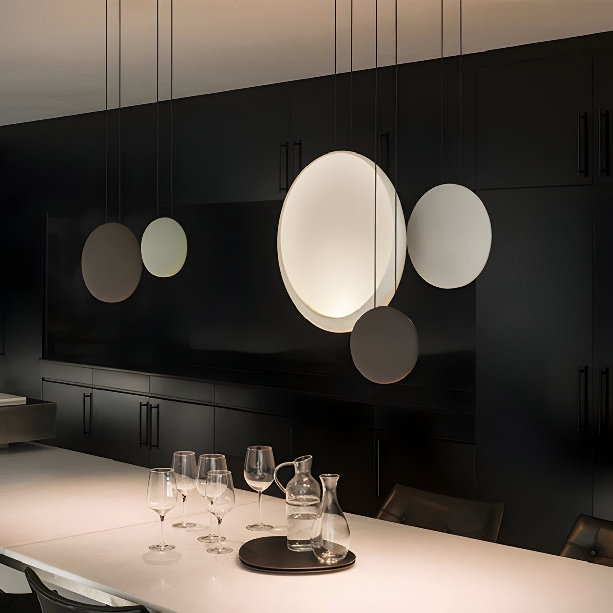 A modern dining area features minimalistic decor with black cabinetry, a sleek Cosmos LED Small Pendant Light, a white table, glassware, and a carafe on a serving tray—creating an elegantly lit, energy-efficient, uncluttered space.