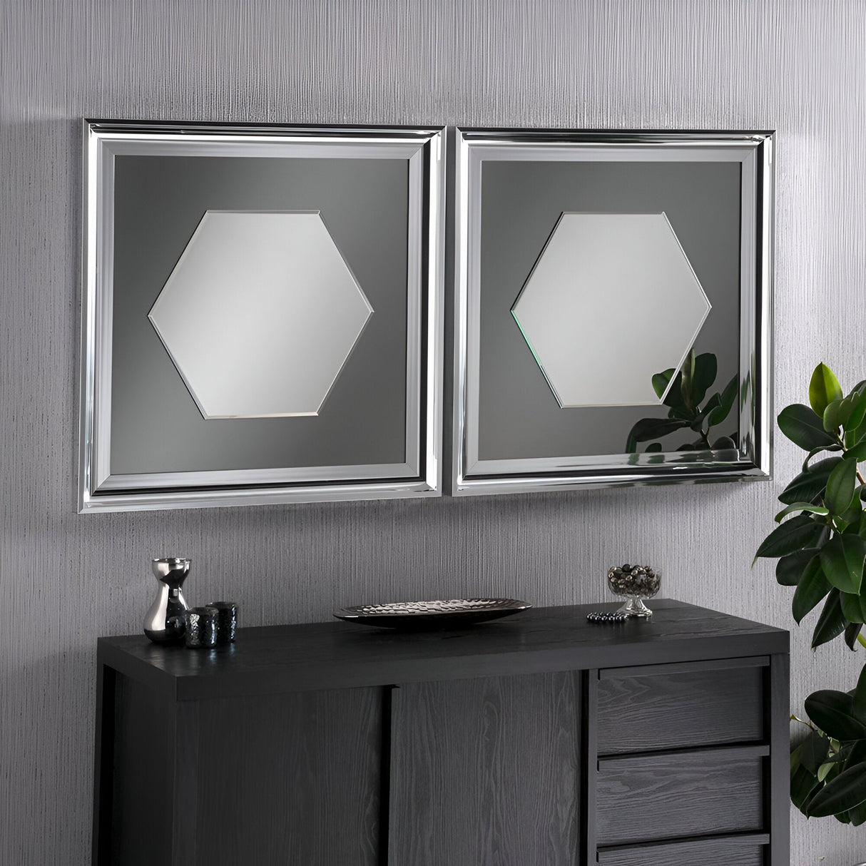 In a contemporary interior, two hexagonal mirrors with square frames are displayed on a textured gray wall, reminiscent of the Croma Rectangle Wall Mirror Grey Lacquered - 74x74cm. Underneath, a dark wooden cabinet holds decorative pieces, and the vibrant green of a plant enlivens the space next to the sleek grey lacquered finish.