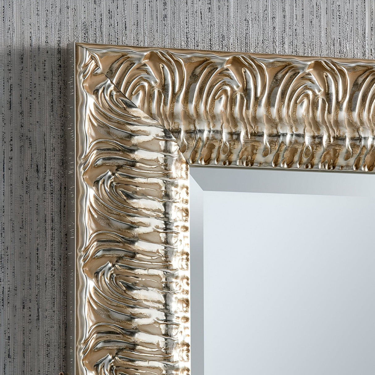 Close-up of an ornate, silver-colored Crown Rectangle Wall Mirror frame with a swirling, textured design. Set against a textured gray wall, its mirror surface reflects light subtly, showcasing intricate details and hinting at modern design elements.