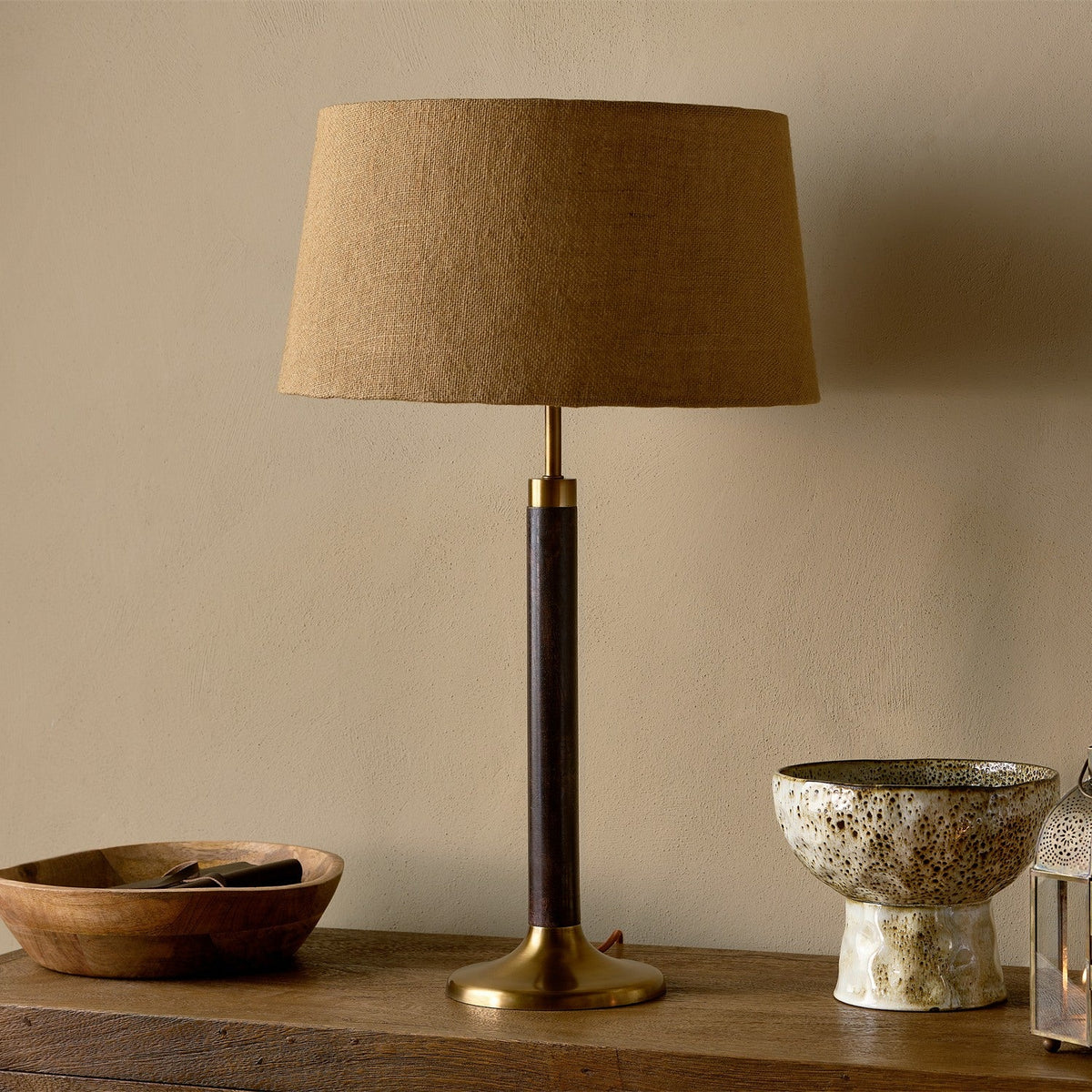 Digha Wooden Table Lamp Base with tan linen drum shade, dark stained mango wood column and antique brass base, shown on a sideboard beside a wooden bowl and ceramic bowl.
