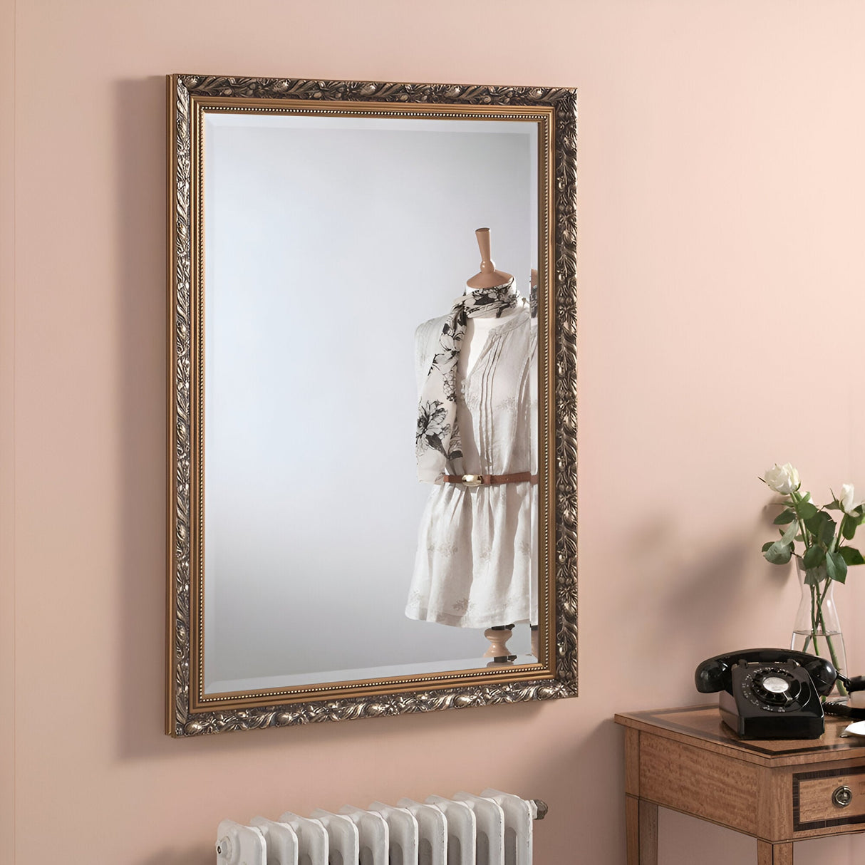 The Dahlia Rectangle Wall Mirror in dark gold, featuring an ornate frame, reflects a dress form adorned with a white top and floral scarf. In the foreground, a wooden table showcases an old-fashioned telephone alongside a vase of white flowers, all set against a light peach wall to evoke vintage-inspired interiors.
