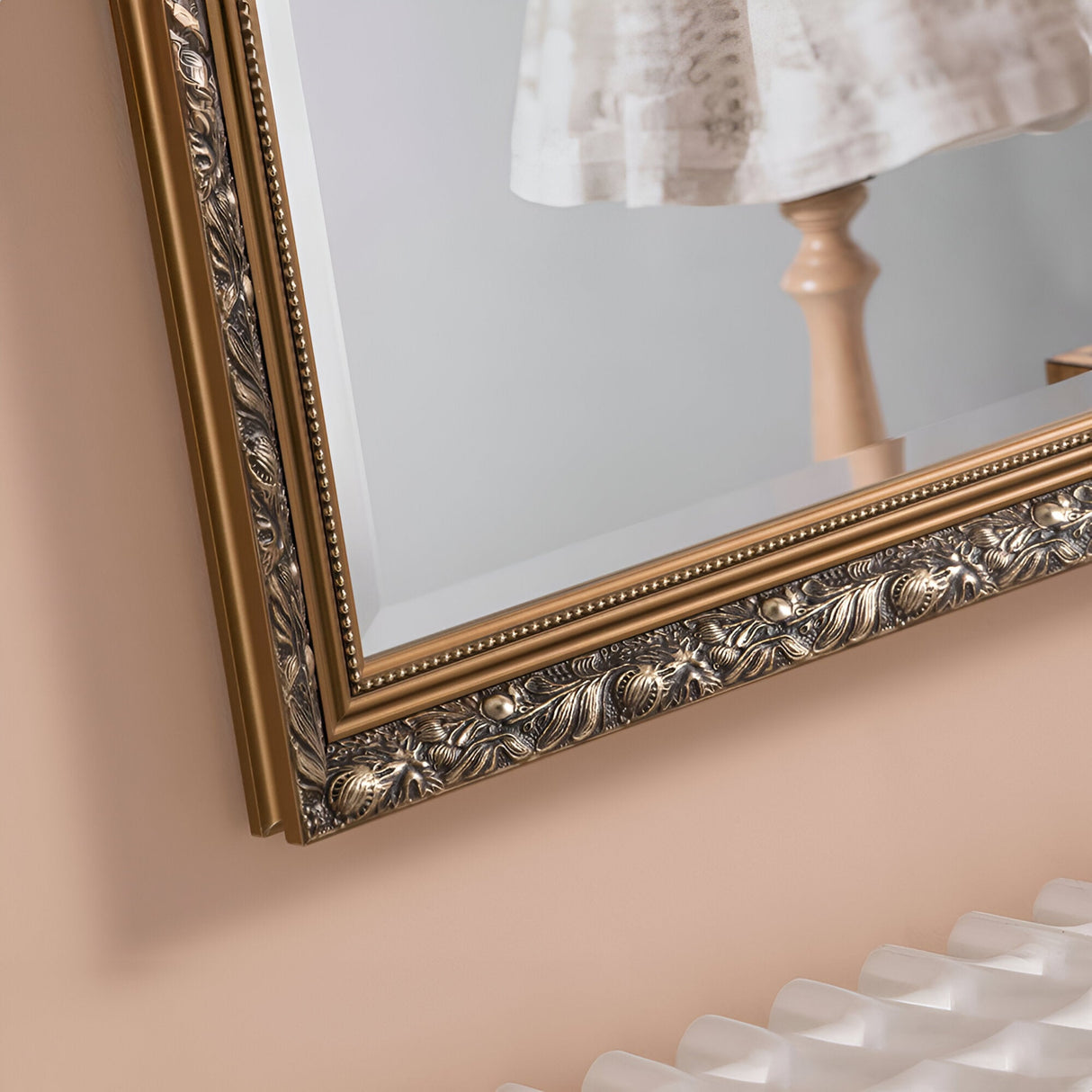 A close-up of the Dahlia Rectangle Wall Mirror in dark gold showcases its intricate floral designs. The reflection reveals a portion of a wooden stand and white lace fabric. The frame is adorned with beaded detailing, making it an ideal addition for vintage-inspired interiors against a warm, neutral-toned background wall.