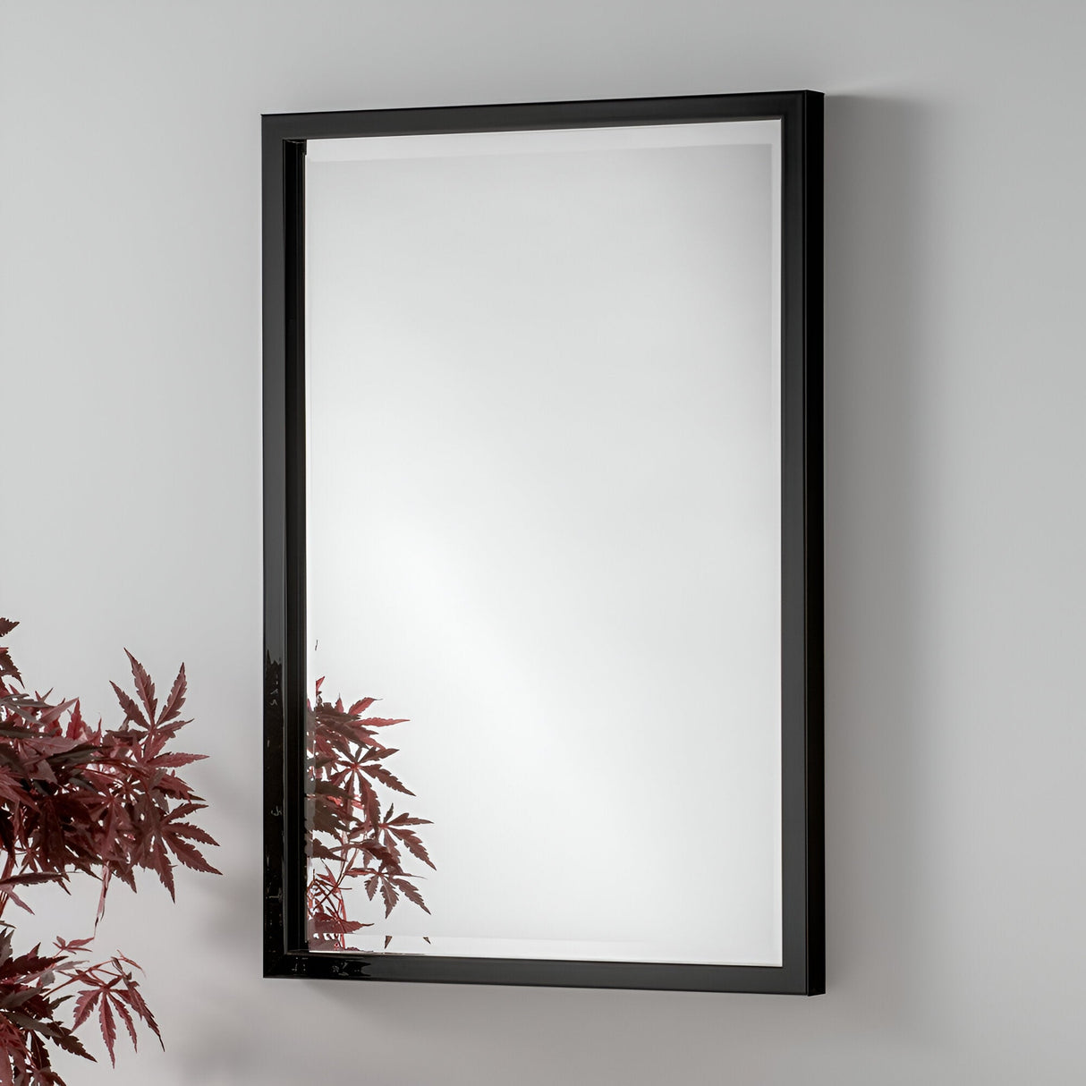 The Dalston Rectangle Wall Mirror - Black is elegantly mounted on a light gray wall. Its sleek frame flawlessly encapsulates the reflection of reddish-hued leaves in the bottom left corner, suggesting nearby greenery—an ideal complement to minimalist interiors.