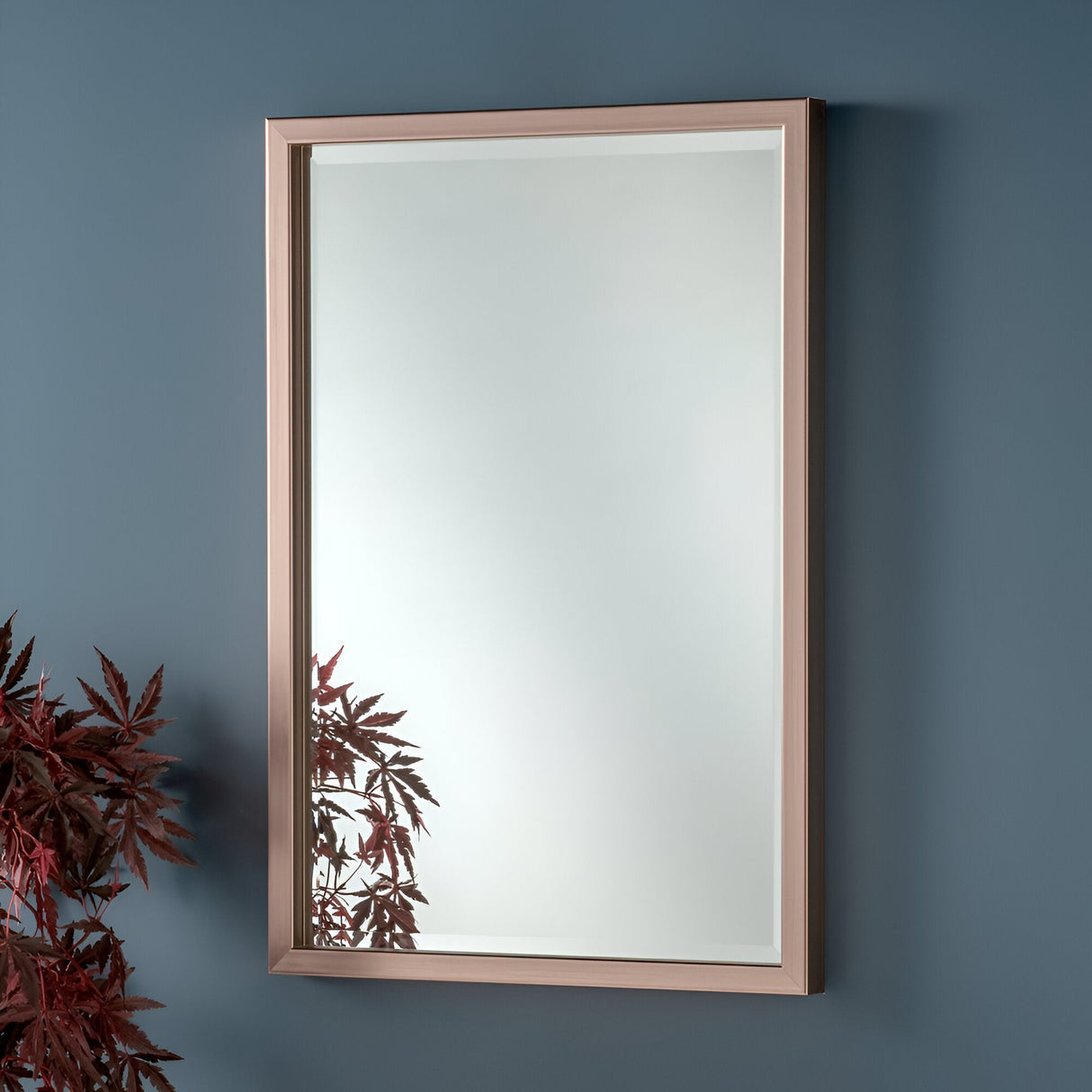 The Dalston Rectangle Wall Mirror - Bronze hangs elegantly on a blue wall, its stylish finish complementing the reflection of red leaves from an offside plant, beautifully integrating this chic decor element with a touch of nature.
