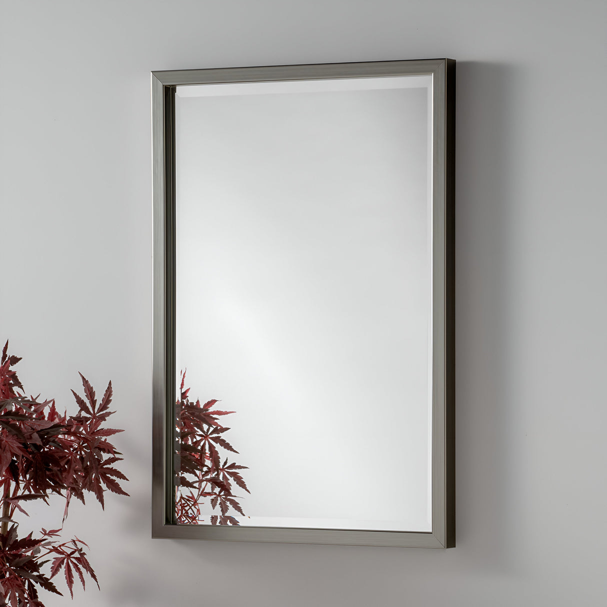The Dalston Rectangle Wall Mirror - Grey, featuring its sleek metallic frame, hangs gracefully on a light gray wall. In the lower left corner, a branch with dark red leaves adds a splash of color and nature to the modern design.