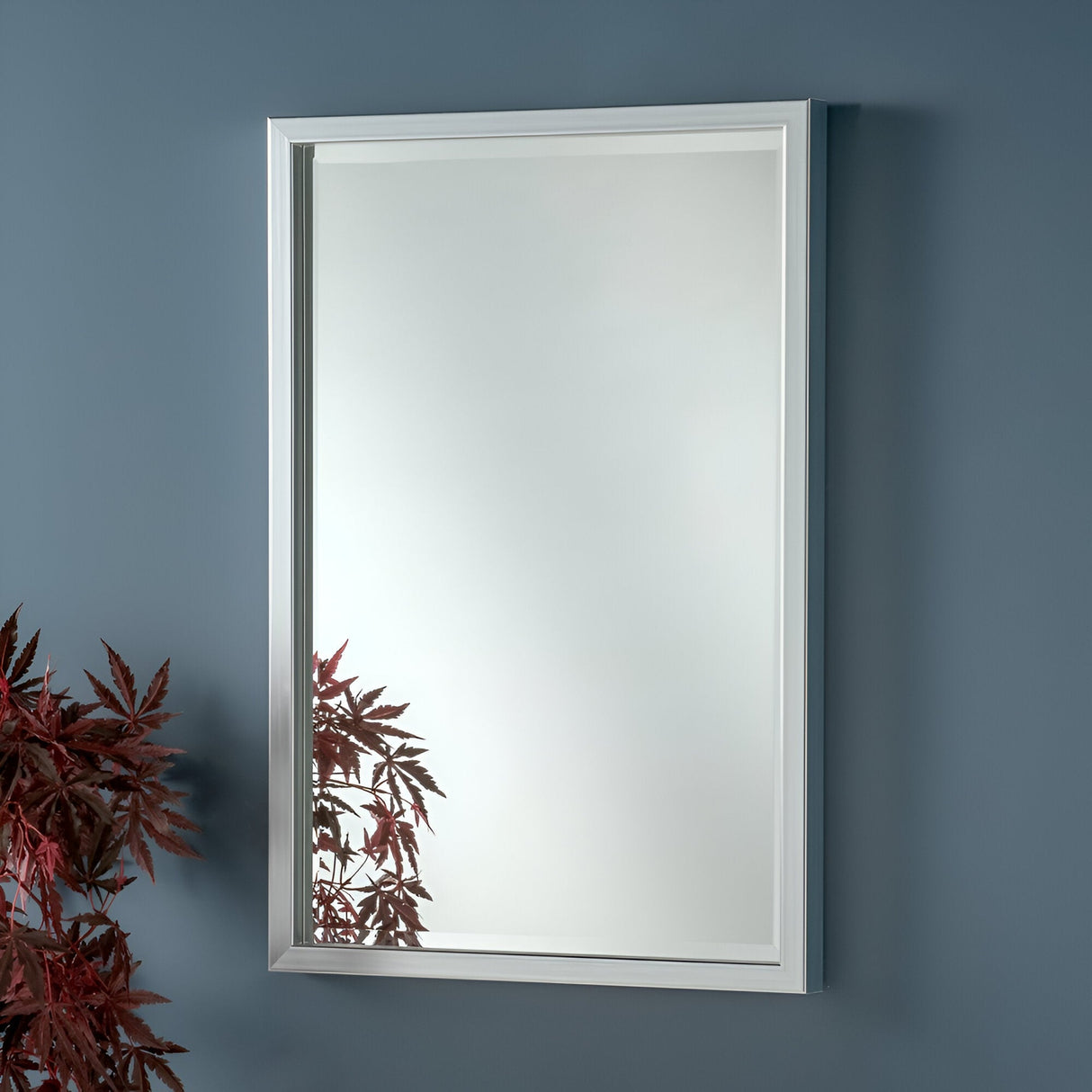 The Dalston Rectangle Wall Mirror - Silver, boasting a sleek frame, is mounted on a blue wall. Its reflection captures red-tinted leaves in the lower corner, infusing colorful accents into the minimalist scene that perfectly complements modern homes.