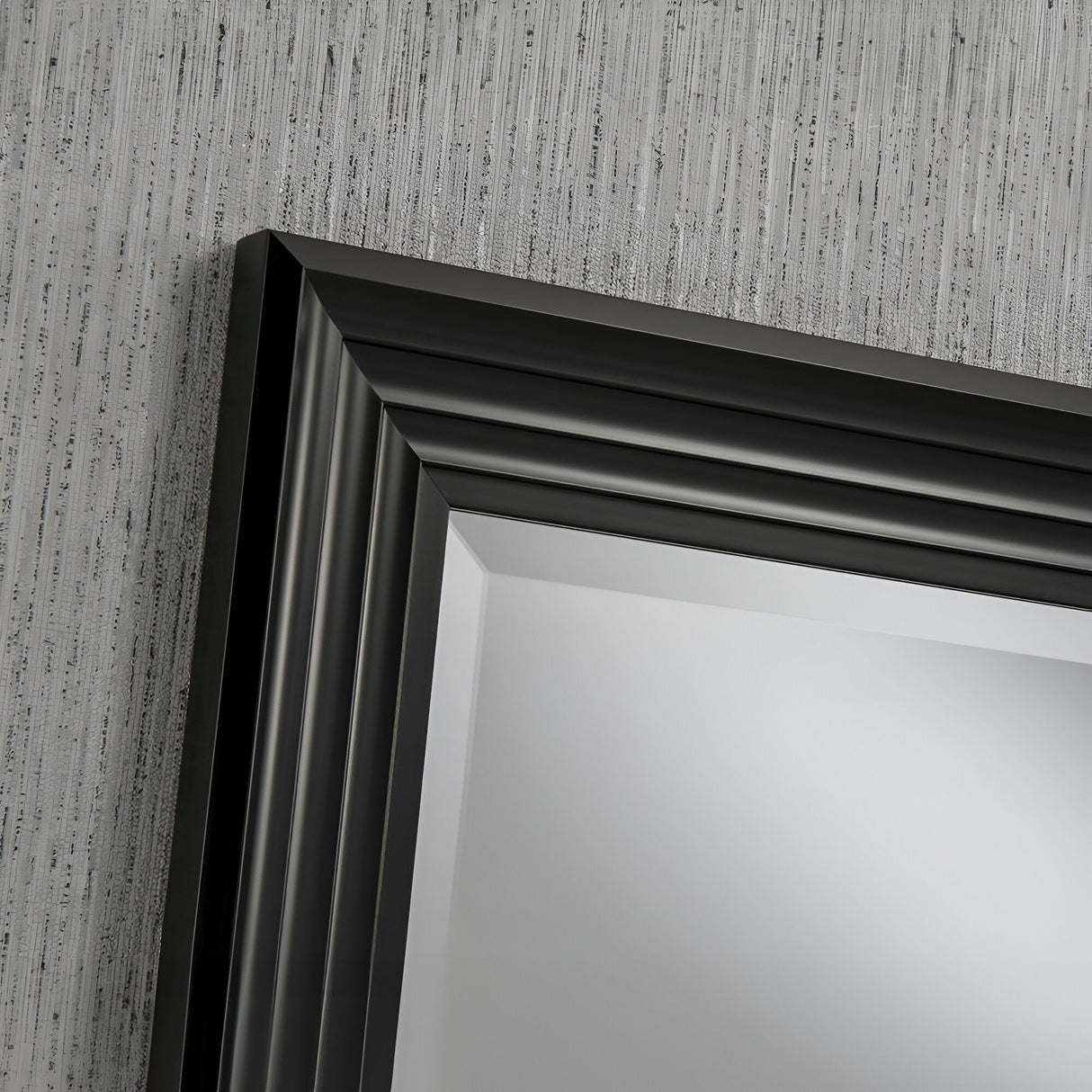The Deco Rectangle Wall Mirror - Black is mounted on a textured gray wall, showcasing its modern design characterized by clean lines and a subtle sheen. The wall's texture offers a sophisticated backdrop, enhancing the mirror's elegant appeal.