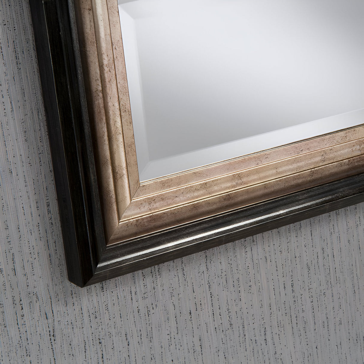 Close-up of a corner of the Deco Rectangle Wall Mirror - Bronze Lacquered with a wide wooden frame featuring metallic and dark tones. This rectangular wall mirror is set against a textured, light-colored wall, adding a touch of elegance to any space.