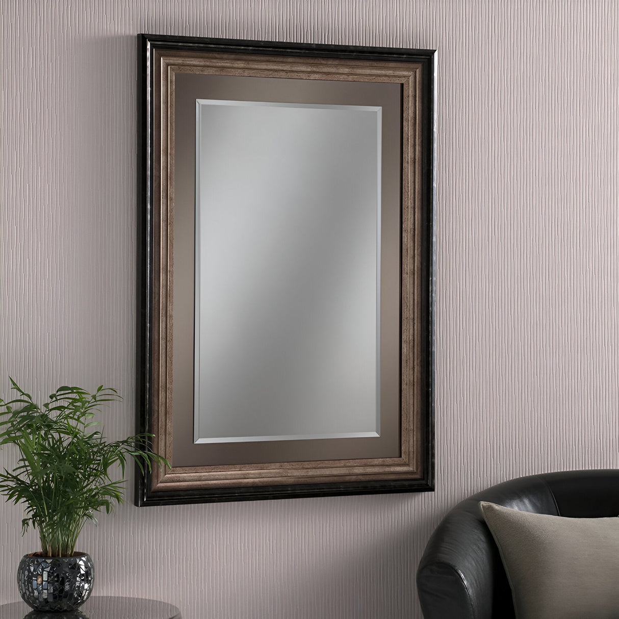 The Deco Rectangle Wall Mirror - Bronze Lacquered hangs on a textured beige wall, with a wide frame. In the corner of a dark leather chair, partially visible at the bottom right, sits a small green plant.