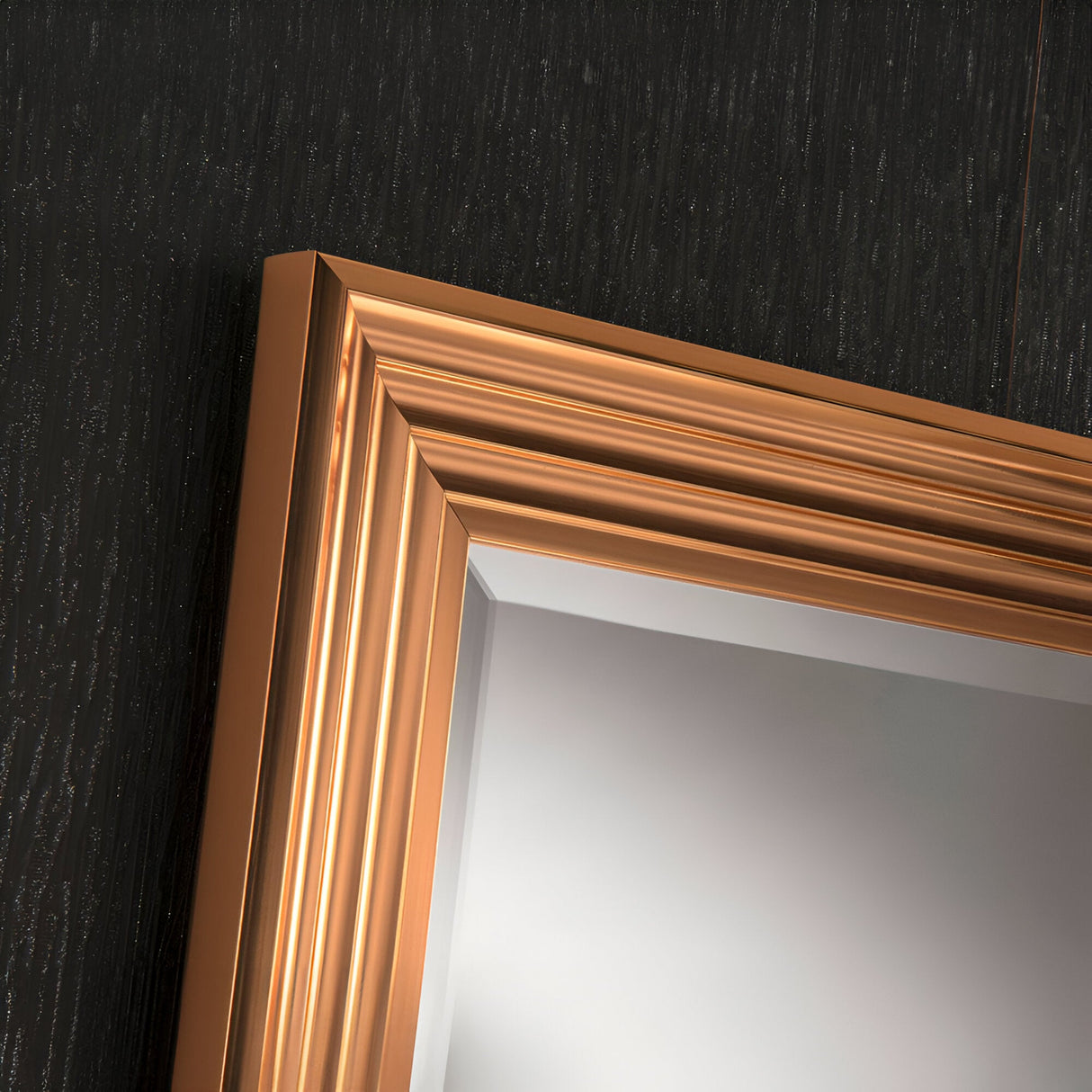 A close-up of the Deco Rectangle Wall Mirror - Copper showcases its shiny, bronze-colored frame with grooved, layered details. This modern design piece stands out against the textured, dark surface in the background and adds a touch of elegance to home decor.