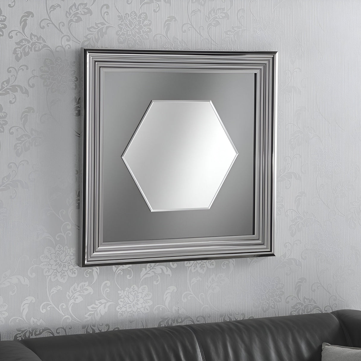 The Opal Hexagon Wall Mirror in Grey Lacquered finish (76x76cm) graces a wall decorated with floral-patterned wallpaper. Beneath this eye-catching mirror, a black leather couch can be seen, seamlessly merging style and elegance.