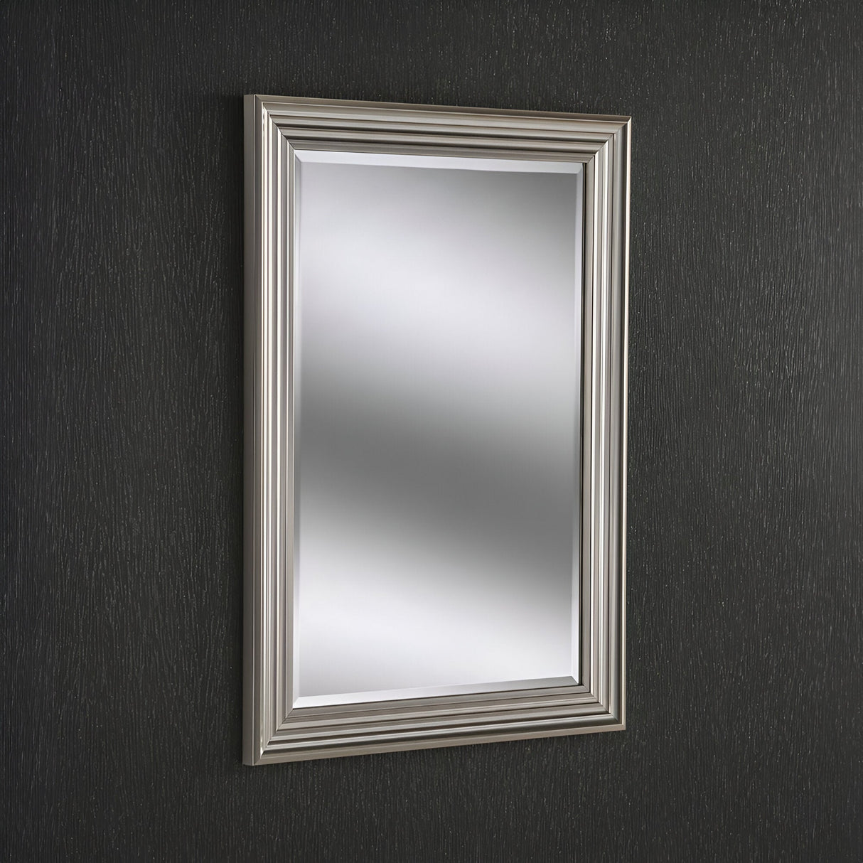 A Deco Rectangle Wall Mirror - Silver is mounted on a dark textured wall. Its smooth, reflective surface embodies modern simplicity, elegantly channeling light throughout the room.