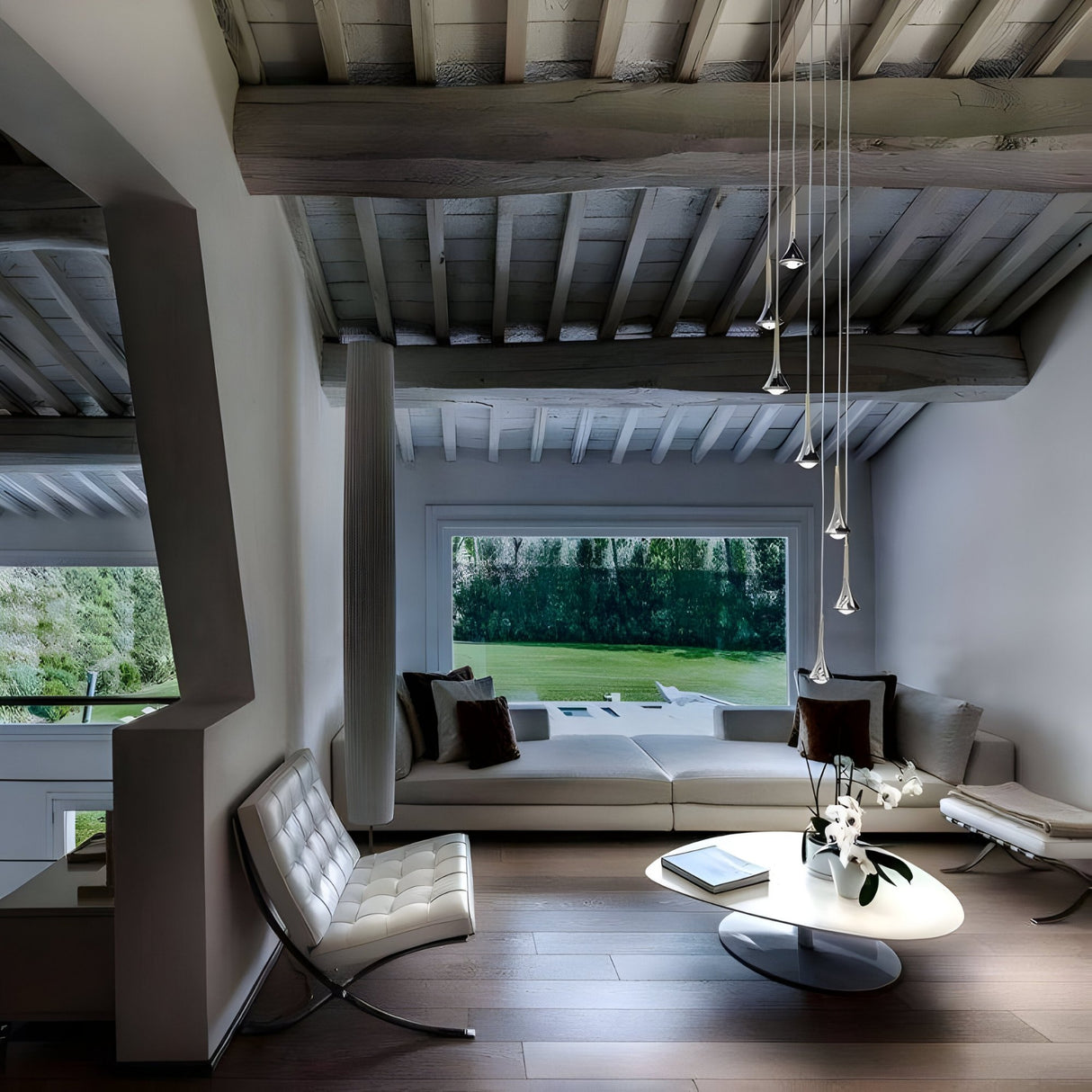 A modern living room with a vaulted wooden ceiling, white furniture, a low coffee table with flowers, the Droplet LED Pendant Light in white & black, and a large window overlooking greenery.