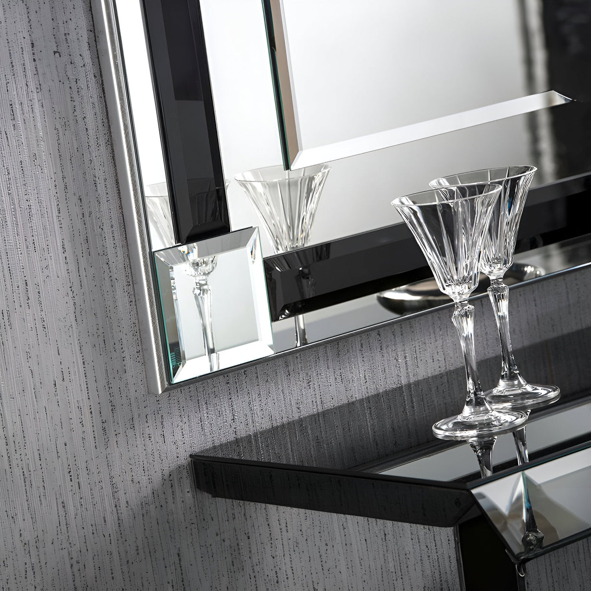 A close-up of an elegant setting showcases two crystal martini glasses resting on a mirrored console table with a contemporary touch. In the background, the Glowis Rectangle Wall Black - 117x84cm mirror, framed in black, is set against a textured metallic silver-gray wall.