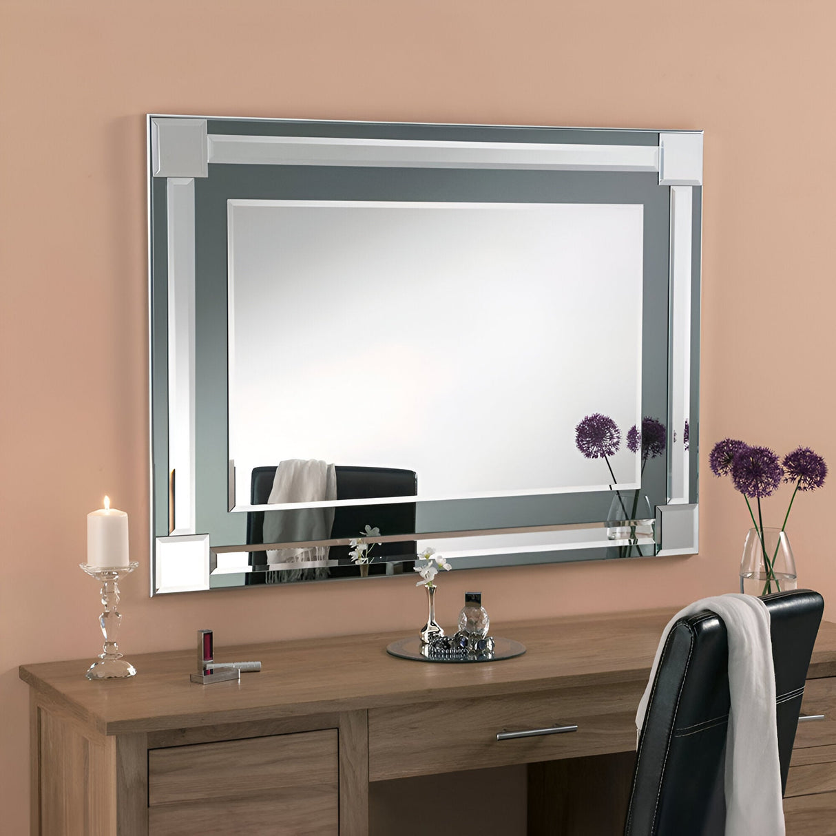 A large Glowis Rectangle Wall Mirror in grey, measuring 117x84cm with beveled edges, hangs above a wooden vanity, infusing the space with modern design. The vanity is complemented by a chair on which a white cloth is draped over the back, along with a candle and two purple flowers elegantly displayed in a vase. The warm beige walls create an inviting backdrop for this minimalist setup.