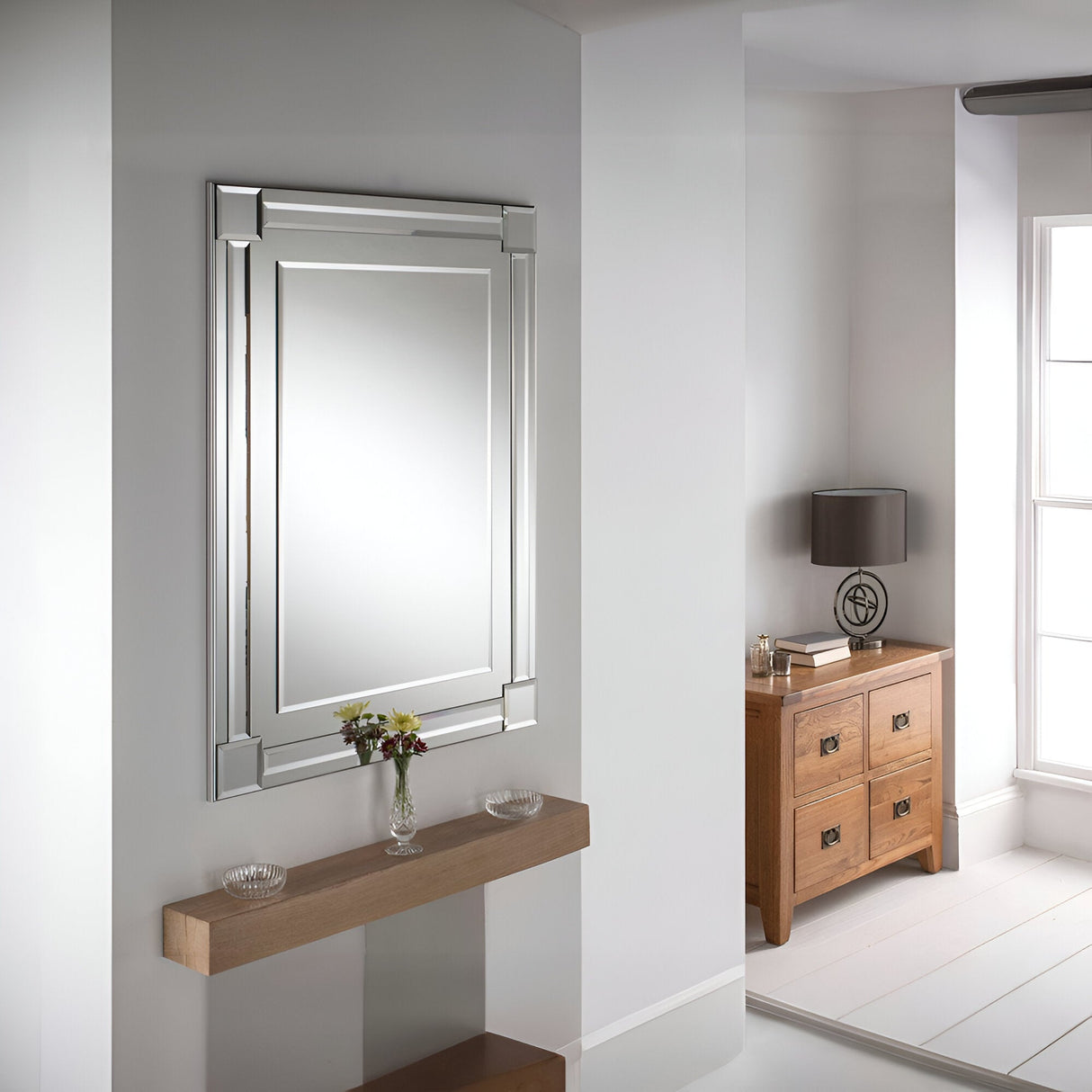 A modern interior showcases the Glowis Rectangle Wall Silver mirror, measuring 117x84cm, prominently displayed on a light gray wall. Below it, a wooden shelf supports a small vase filled with flowers. To the right, a wooden dresser is adorned with a lamp and various decorative items, while natural light floods in through the window, capturing the essence of minimalist design.