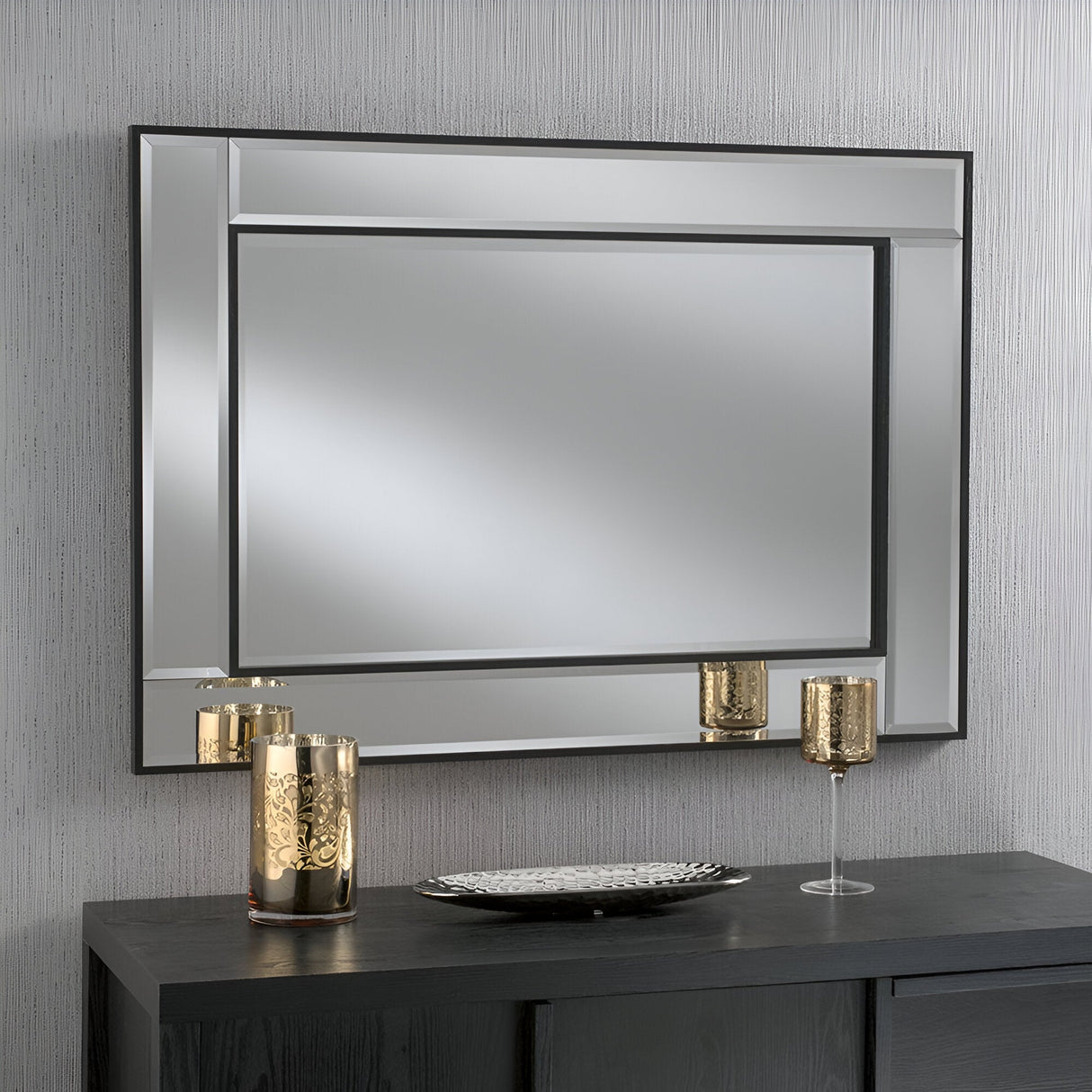 The Venire Rectangle Wall Mirror Black - 106x75cm, with its geometric frame, hangs on a light-textured wall, creating an atmosphere that feels spacious and inviting. Below it, a black console table gracefully holds two gold candle holders alongside a stylish silver tray, exuding a sense of minimalistic elegance.