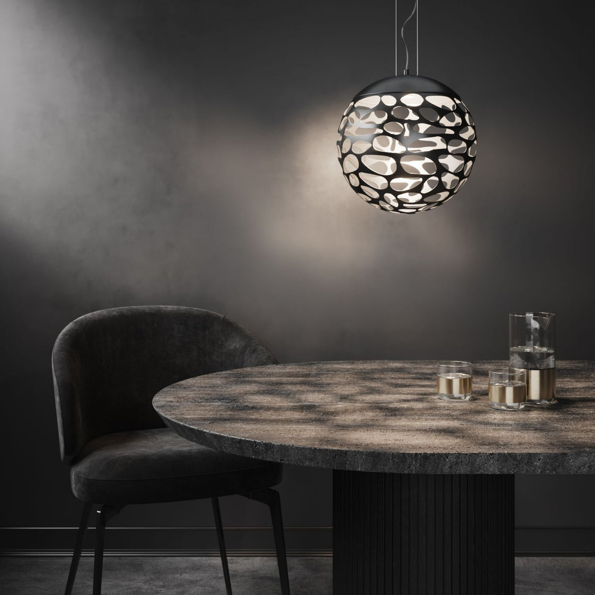 A modern dining area featuring a round stone table, a dark matte black upholstered chair, and the Elon LED Light Pendant - Matte Black casting patterned shadows; two glasses and a glass of water rest on the table.