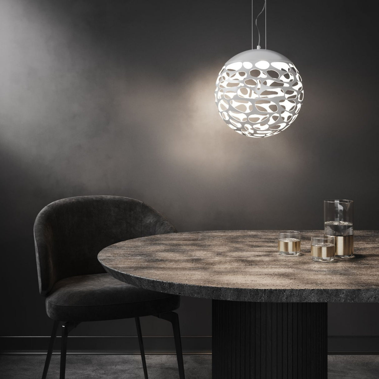 A modern dining area features a round stone table, two glasses and a carafe, a dark upholstered chair, and an Elon LED Light Pendant in Matte White with cut-out patterns that cast intricate ambient shadows on the dark wall.