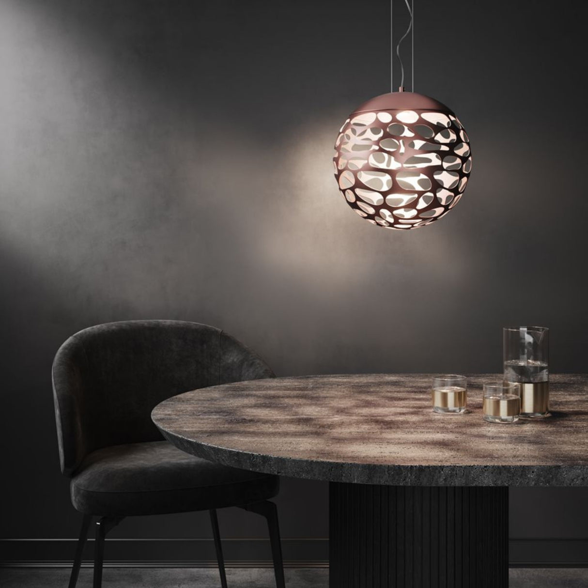 A modern dining area features a round, textured table, two glasses with a carafe, a dark upholstered chair, and the Elon LED Single Pendant - Copper casting patterned illumination on the gray wall.