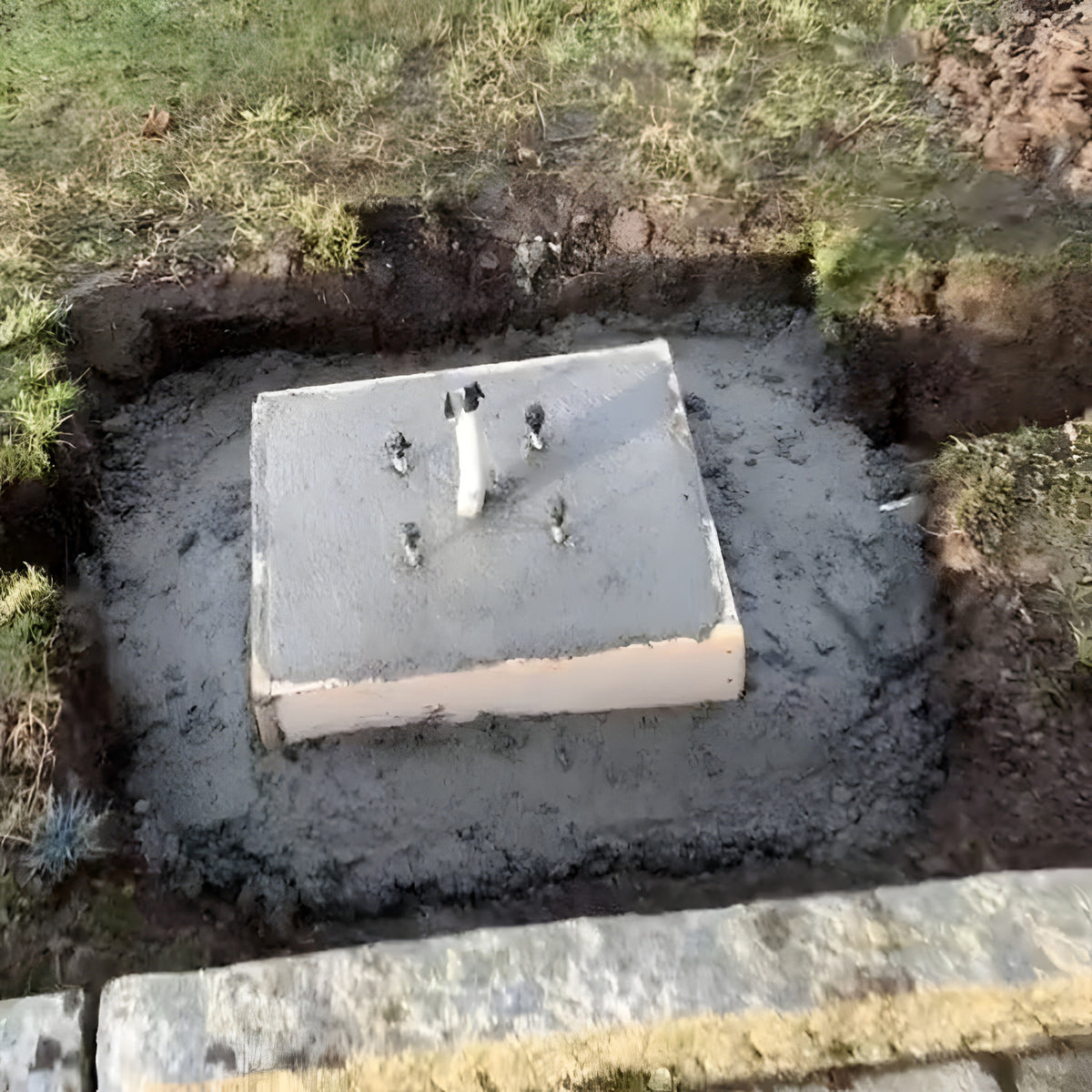 Medium Foundation Frame 150mm Square set in wet concrete outdoors, showing four galvanised steel studs and a central conduit pipe within a dug garden trench.