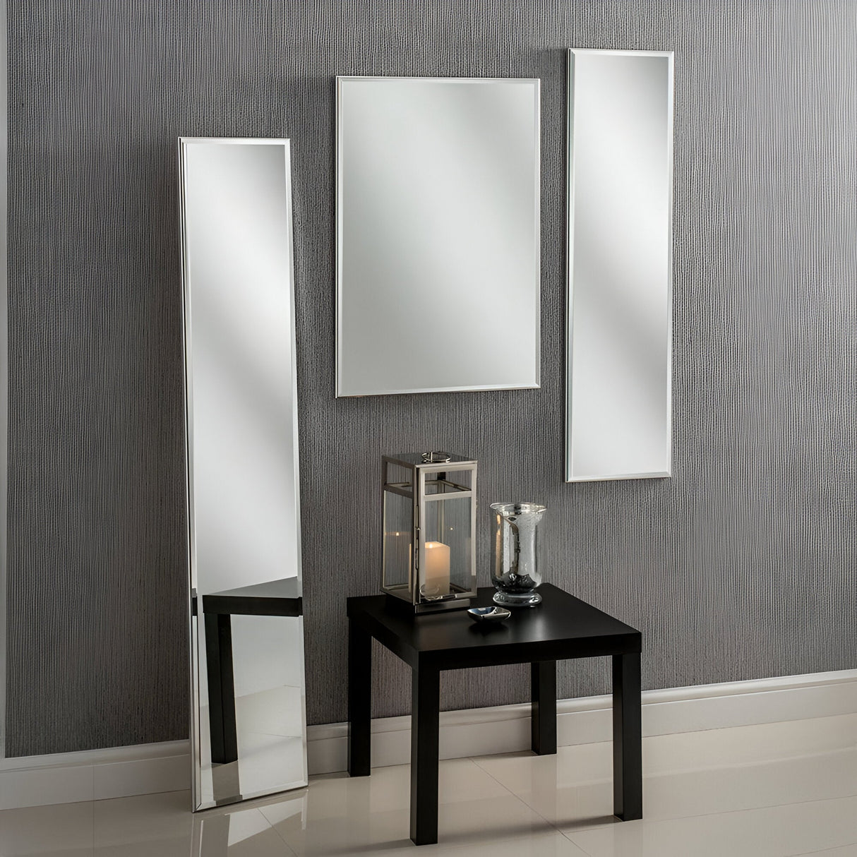 A modern interior displays three Avante Rectangle Wall Mirrors with minimalist silver wood frames on a textured gray wall. Below them, a black table supports a lantern and a small vase. The design is clean and minimalist, enhanced by beige tiled flooring.