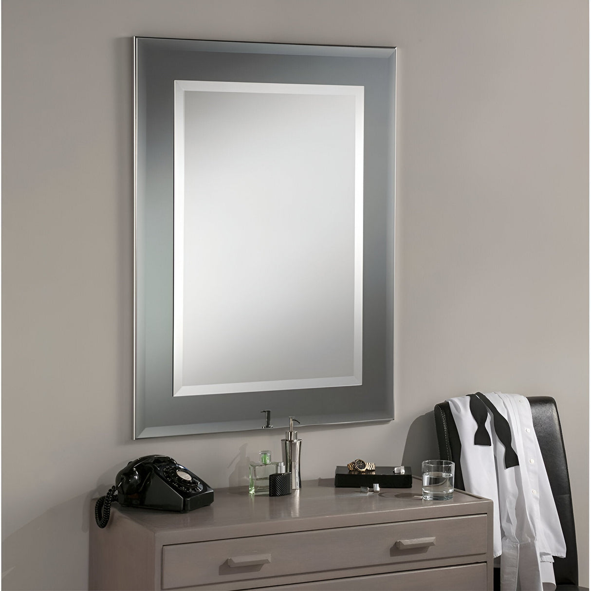 A sleek, modern bathroom features the Vivid Rectangle Wall Mirror - Grey with its minimalist design above a beige vanity. On the counter sits a vintage black rotary phone, alongside a glass of water and a neatly folded white and black shirt, adding an elegant touch to the contemporary decor.