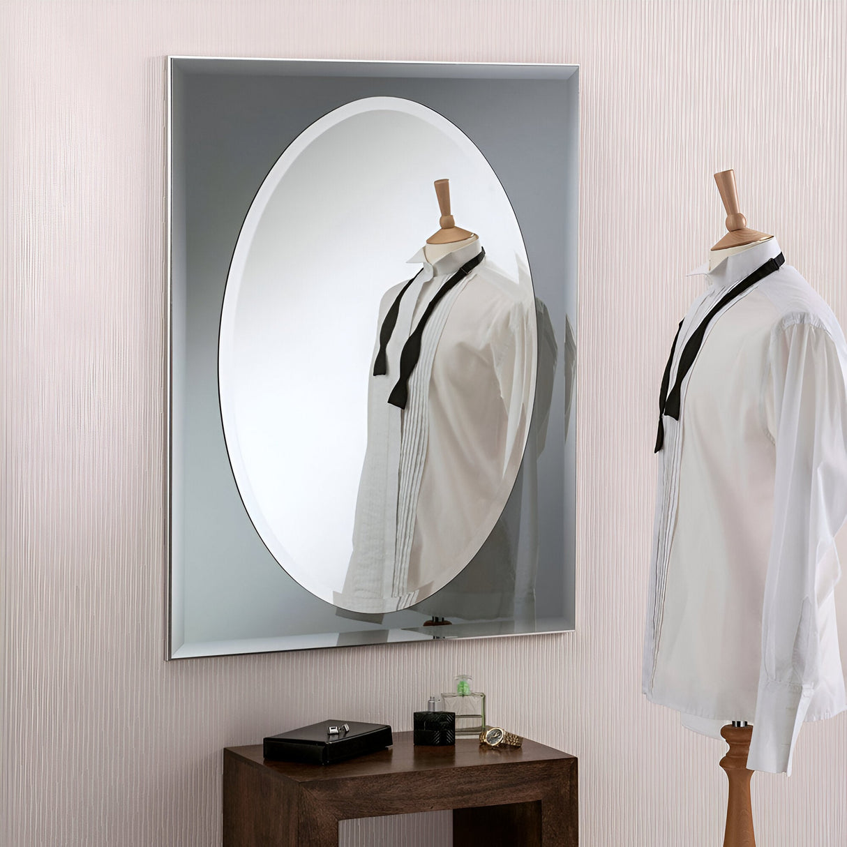 A wooden mannequin dressed in a white shirt and black tie stands in front of the Vivid Oval Wall Mirror Grey - 83x62cm. The mirror's reflection enhances the scene with a decorative touch. Beneath it, a wooden table displays a small black box and a green perfume bottle.