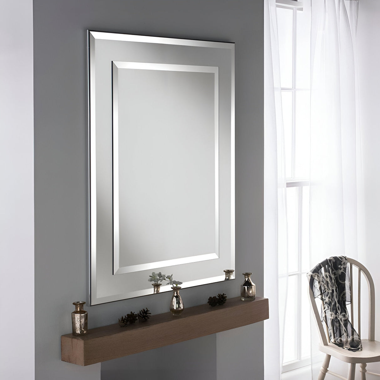 The Vivid Rectangle Wall Mirror - Silver is elegantly displayed on a gray wall. Beneath it, a wooden shelf is beautifully adorned with decorative candles and a small plant. Nearby, a white chair draped with a patterned scarf sits beside a window dressed with sheer curtains, enhancing the modern interior aesthetic.
