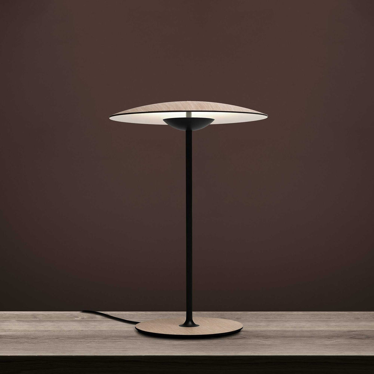 The Ginger LED Table Lamp - Black & Oak features a slim stand and wide circular shade, shown illuminated on a wooden surface against a plain brown background, softly lighting the area.