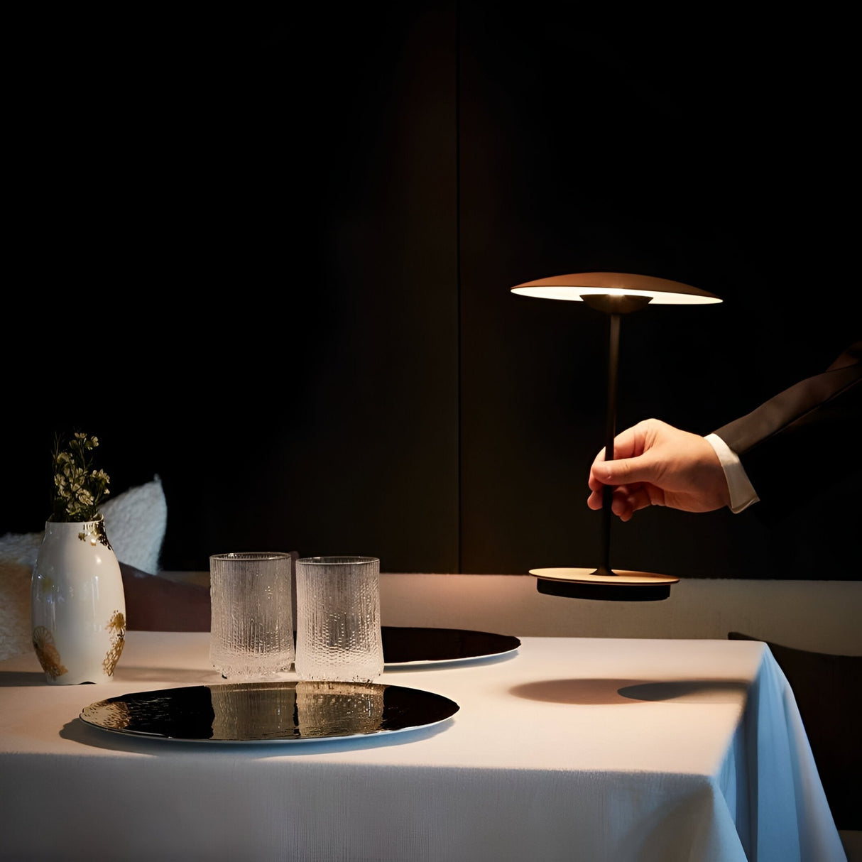 A hand adjusts the Ginger LED Table Lamp in Black & Oak on a white table with two textured glasses, a reflective tray, and a white vase of small flowers, showcasing modern sophistication against a dark backdrop.