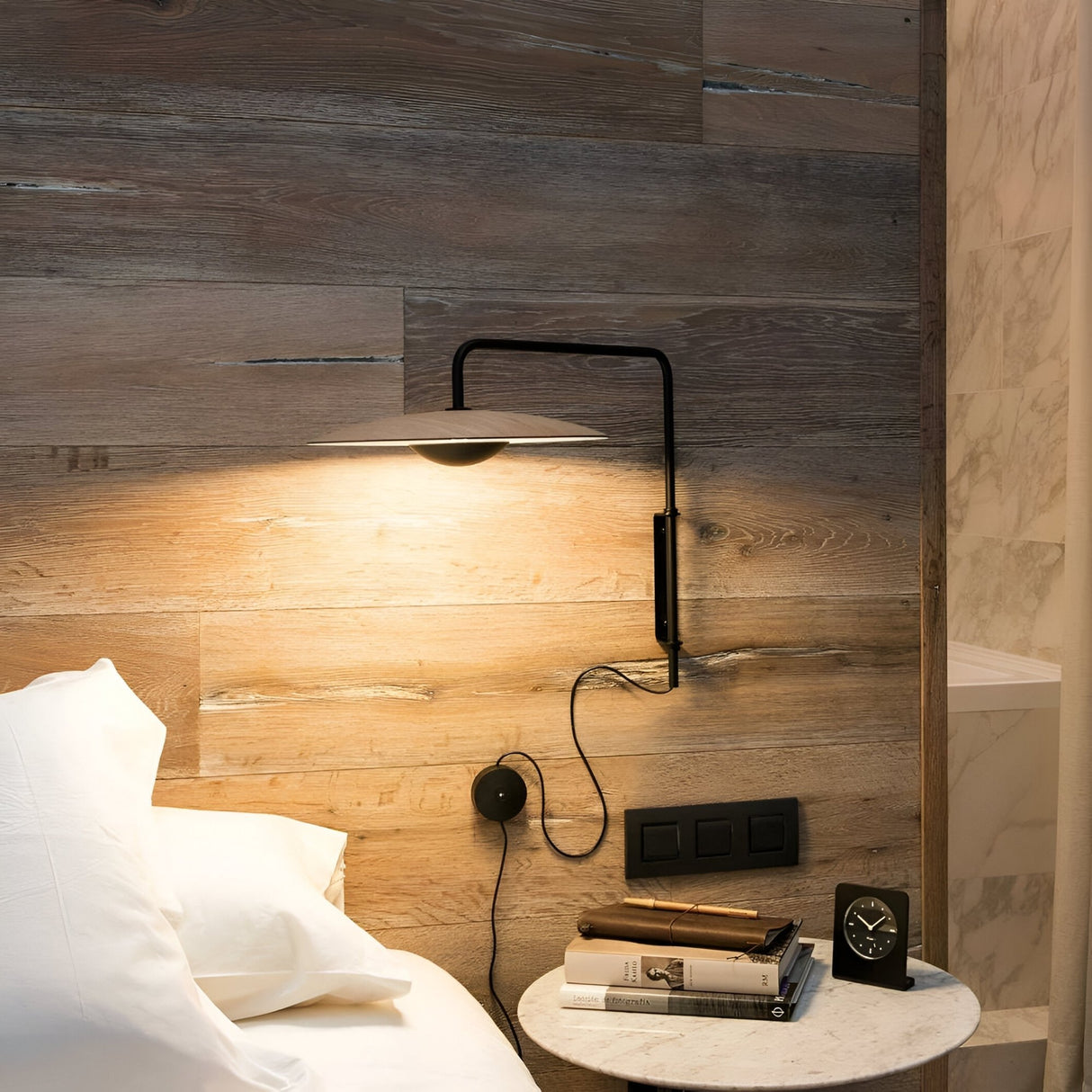 A cozy bedroom features the Ginger LED Wall Light - Black & Oak illuminating a wooden headboard, with a white pillow on the bed and a round marble nightstand holding books, a clock, and glasses.
