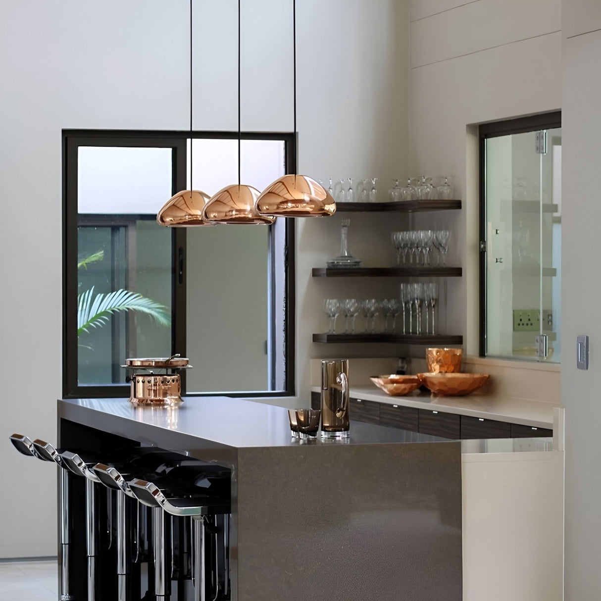 A modern kitchen features a gray island, four black barstools, the Gulf Glass Pendant Light - Copper, glass shelves with glasses, a window, and metallic kitchenware. Neutral tones and minimalistic design create an inviting atmosphere.