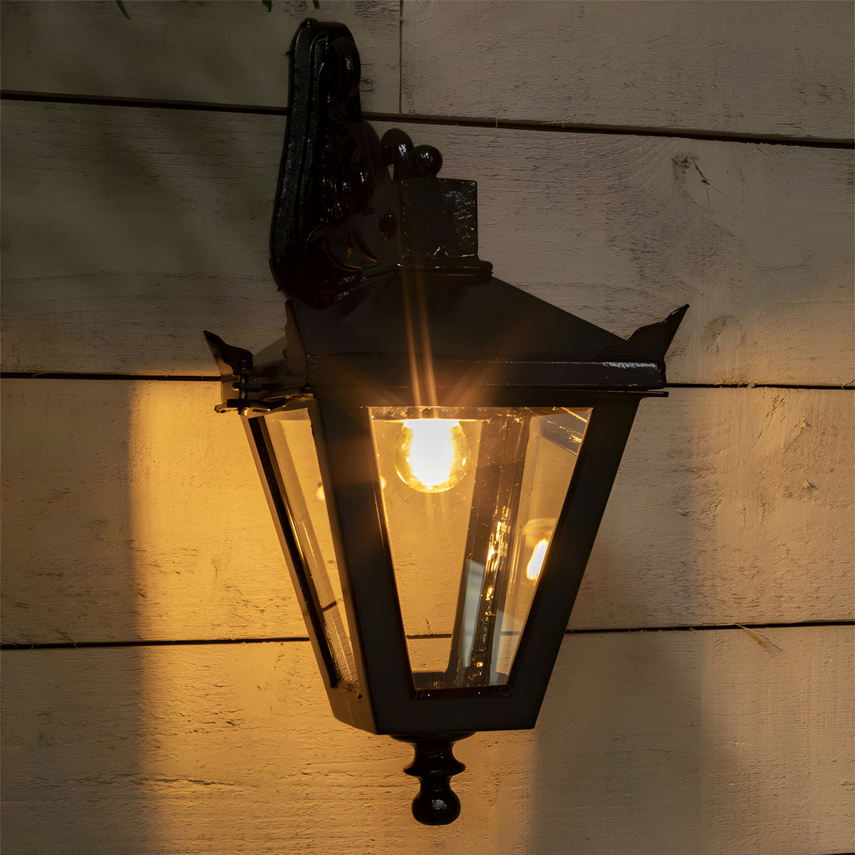 Victorian Cast Iron Downturned Wall Lantern in black, tapered four-sided glass head with ornate mounting bracket, lit and mounted on a wooden panelled wall.
