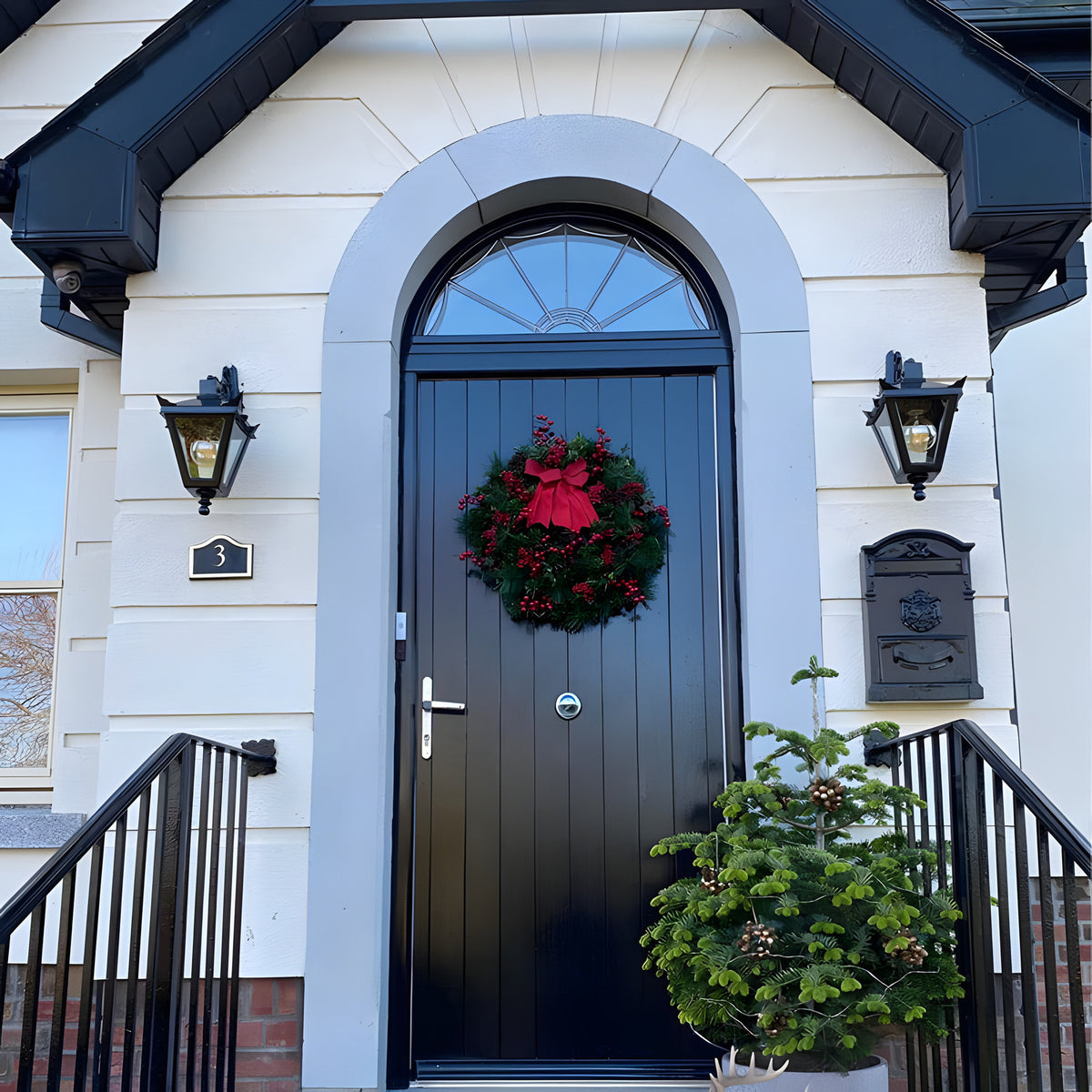 Black cast iron downturned wall lantern with tapered four-sided glass head, stepped crown and corner finials, mounted beside a black front door on a white house.