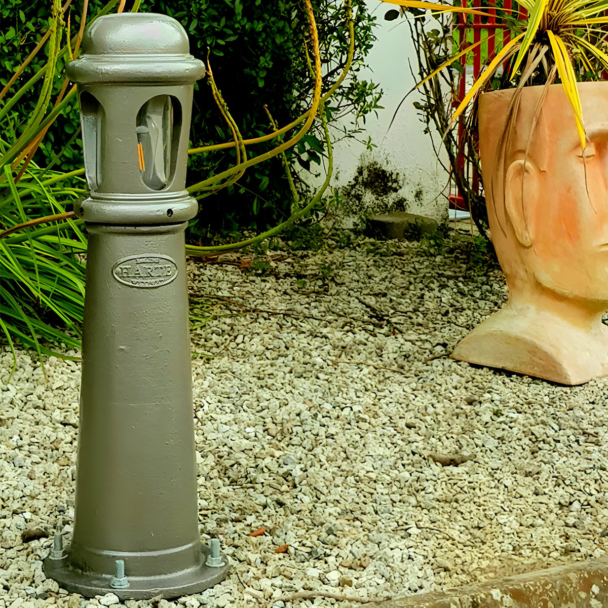 Medium Victorian cast iron bollard light in a grey-green finish with frosted lens window, base fixing bolts, set in gravel garden with plants and terracotta planter.