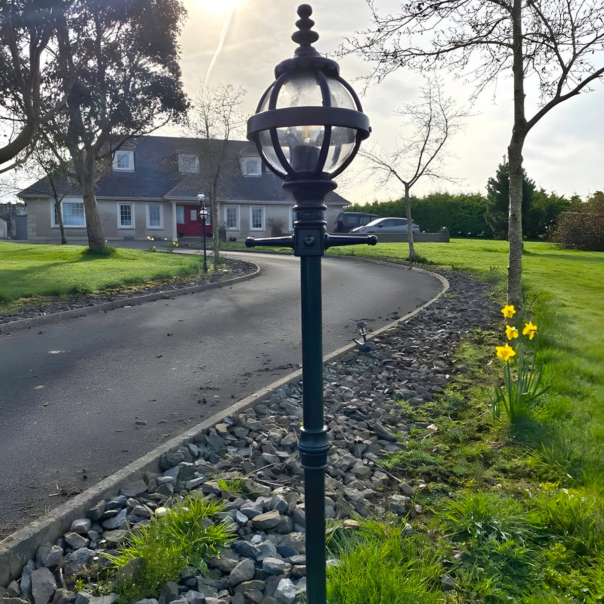 Victorian Cast Iron Globe Lamp Post 1.4m in black, with a caged globe shade on a decorative pole, installed on a gravel border beside a driveway.
