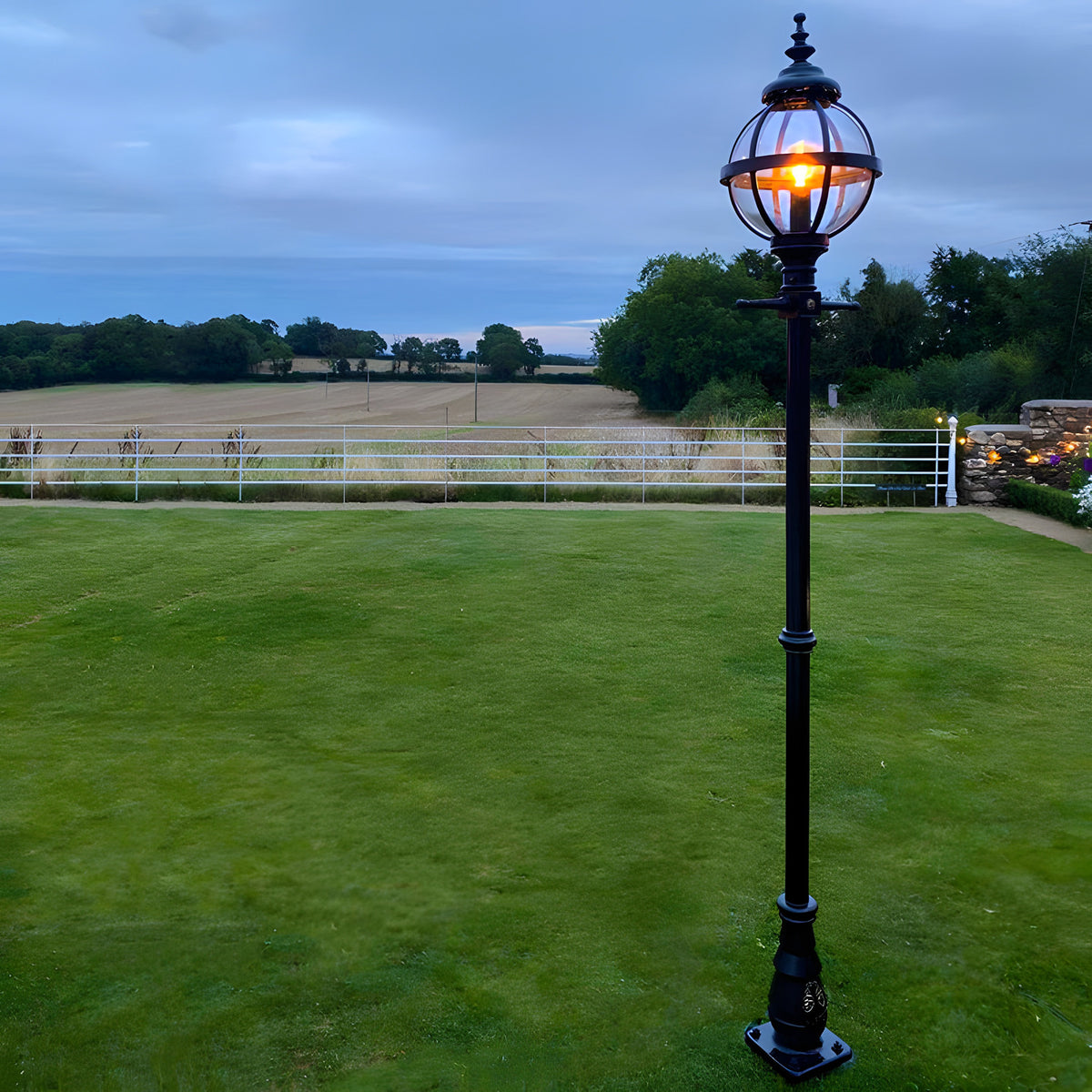 Victorian Cast Iron Globe Lamp Post in black, 2.5m tall with a banded cage globe lit against a dusk garden setting, mounted on a decorative base with a lawn and countryside view behind.