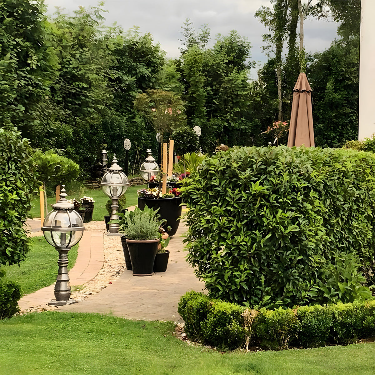 Large Victorian cast iron globe pedestal post light in black, shown installed along a garden pathway edge with clipped hedges and planted pots.