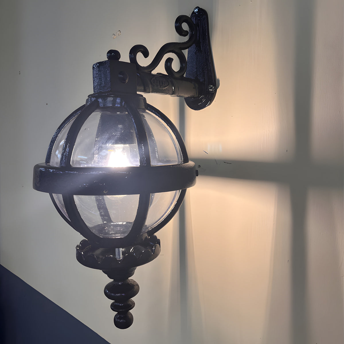 Small Victorian cast iron downturned globe wall lantern in black, with a scrolled wall bracket, banded cage, and polycarbonate globe shade, lit against a plain wall.