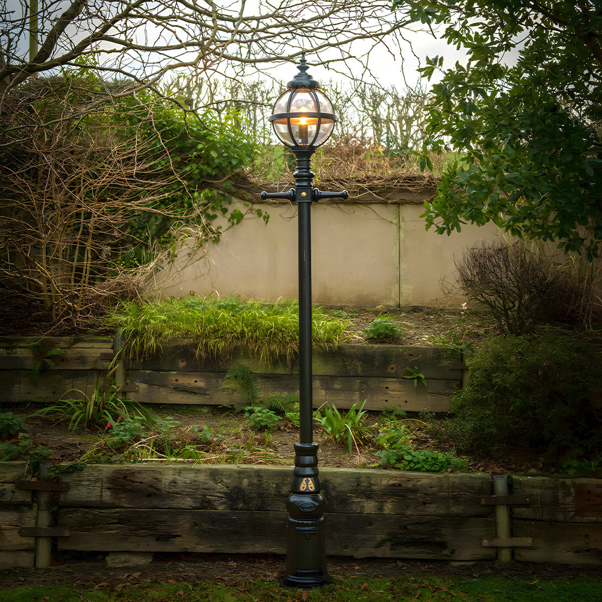 Victorian Manor 3m cast iron globe lamp post in black, lit globe lantern in banded cage atop a tall column, set in a garden against a stone wall and greenery.