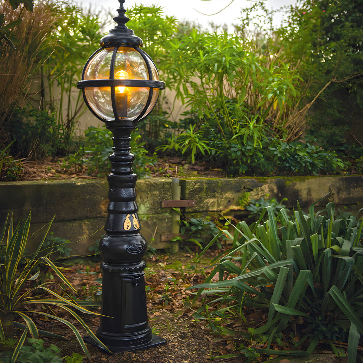 Victorian Manor cast iron globe pedestal post light in black, banded lantern cage on a tall traditional column, shown lit in a garden setting.