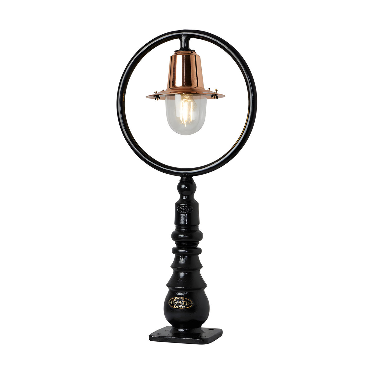 Small railway-style cast iron pedestal post light with a black circular hoop frame, copper spun shade over a clear glass dome, ribbed black base, on a plain white background.