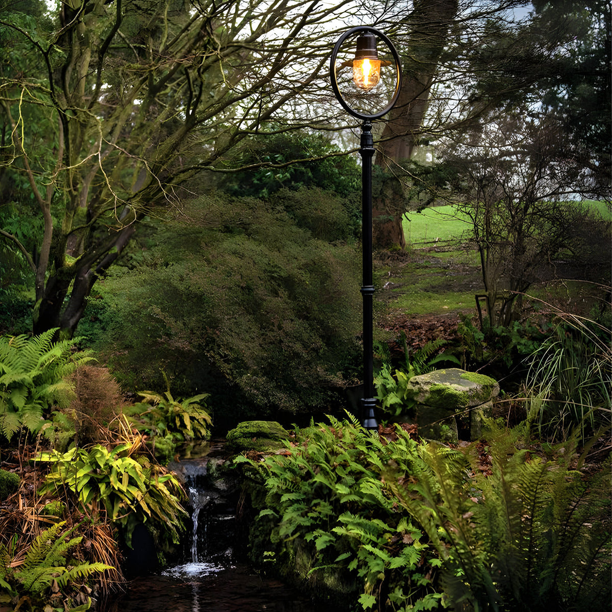 Railway Style Cast Iron Lamp Post in black, 2.43m tall with a lit lantern head under a hoop, set in a lush garden beside a small stream.